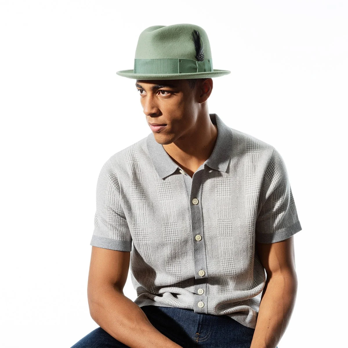 A young man wearing a light grey short-sleeve button-up shirt and a green fedora hat with a black feather, sitting against a white background.