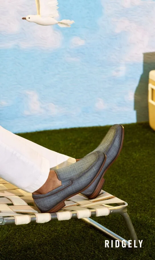 Person wearing gray shoes and white pants resting on a lounge chair outdoors, with a blue sky painted on the wall and a decorative flying bird in the background.