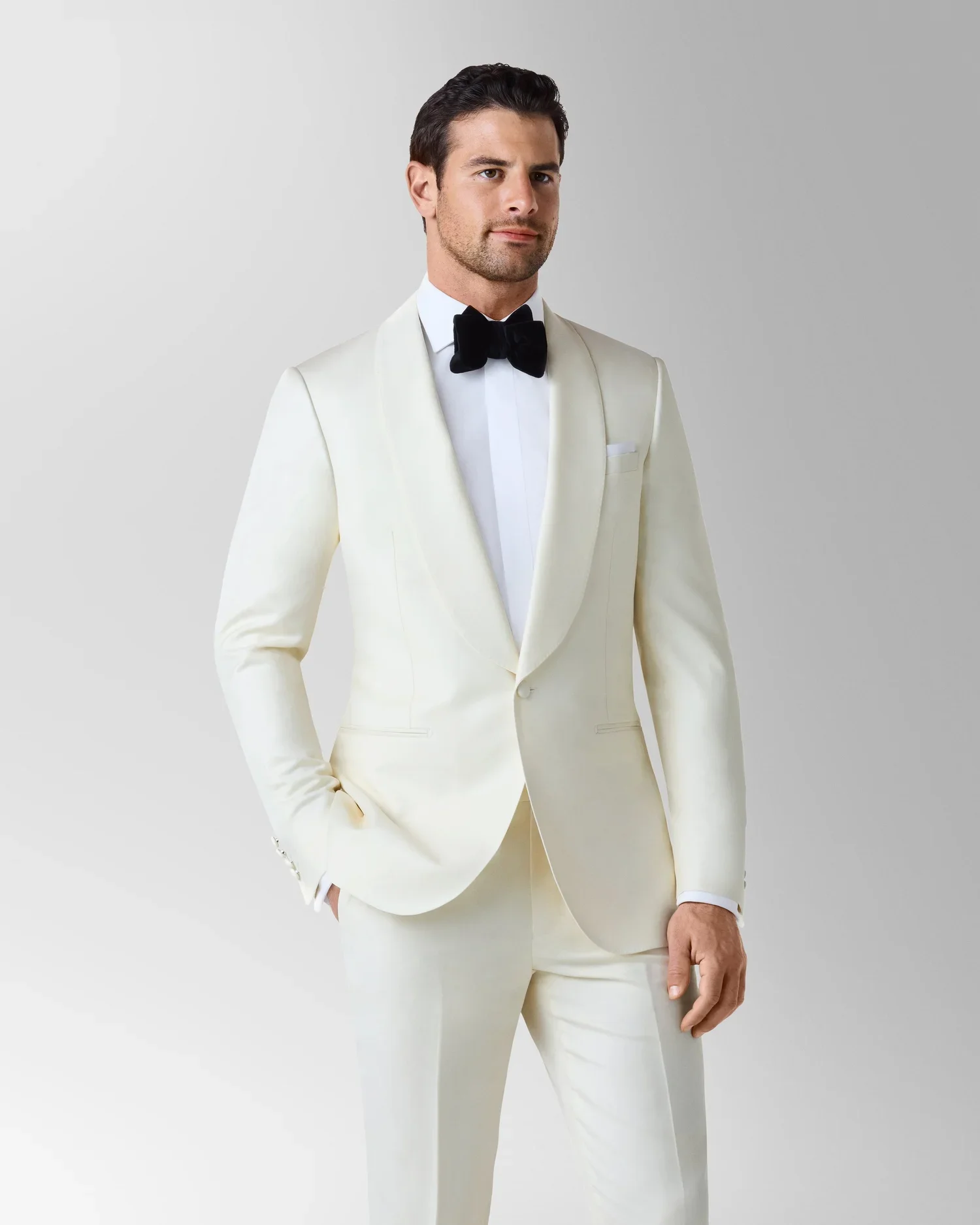 A man dressed in a white tuxedo with a black bow tie, standing against a plain gray background.