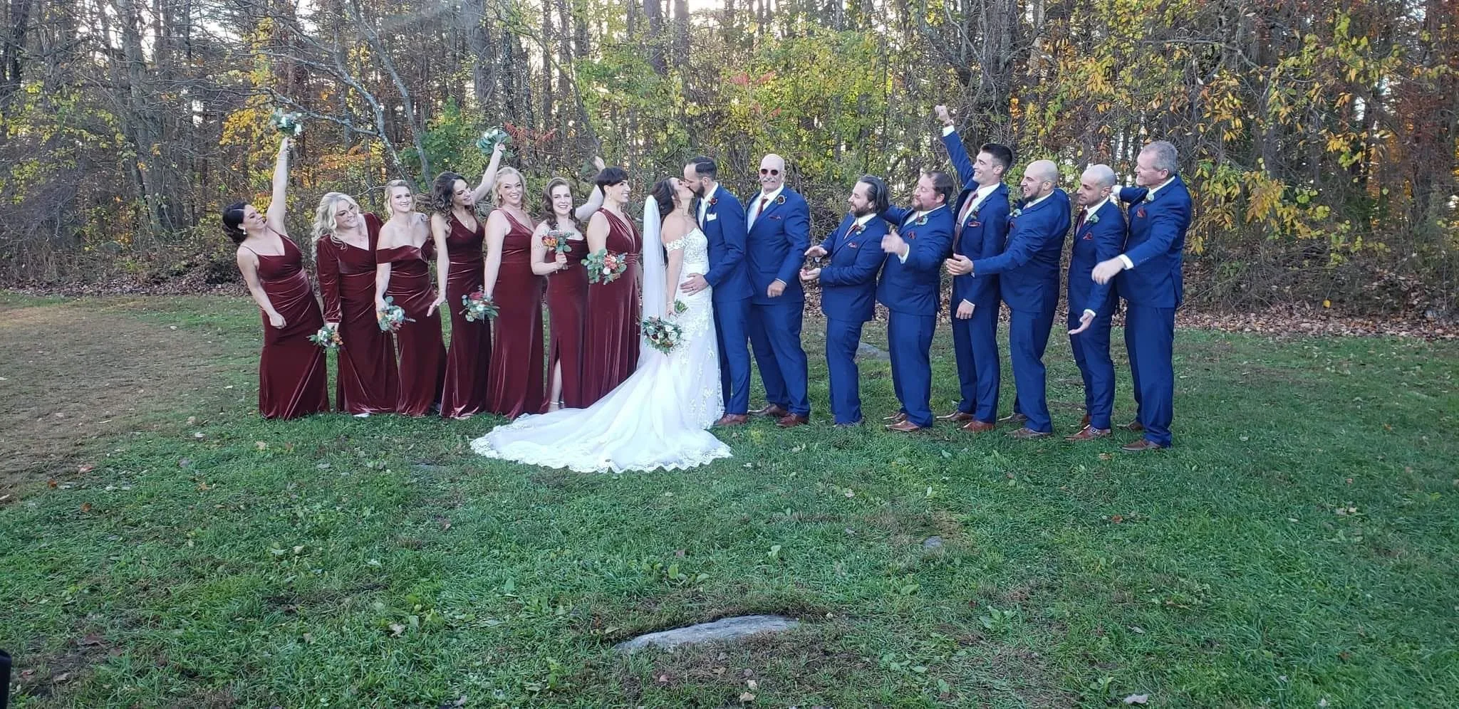 Group of wedding party members outdoors, including the bride and groom at the center, surrounded by bridesmaids in burgundy dresses and groomsmen in blue suits. They are celebrating with some raising their hands and smiling.