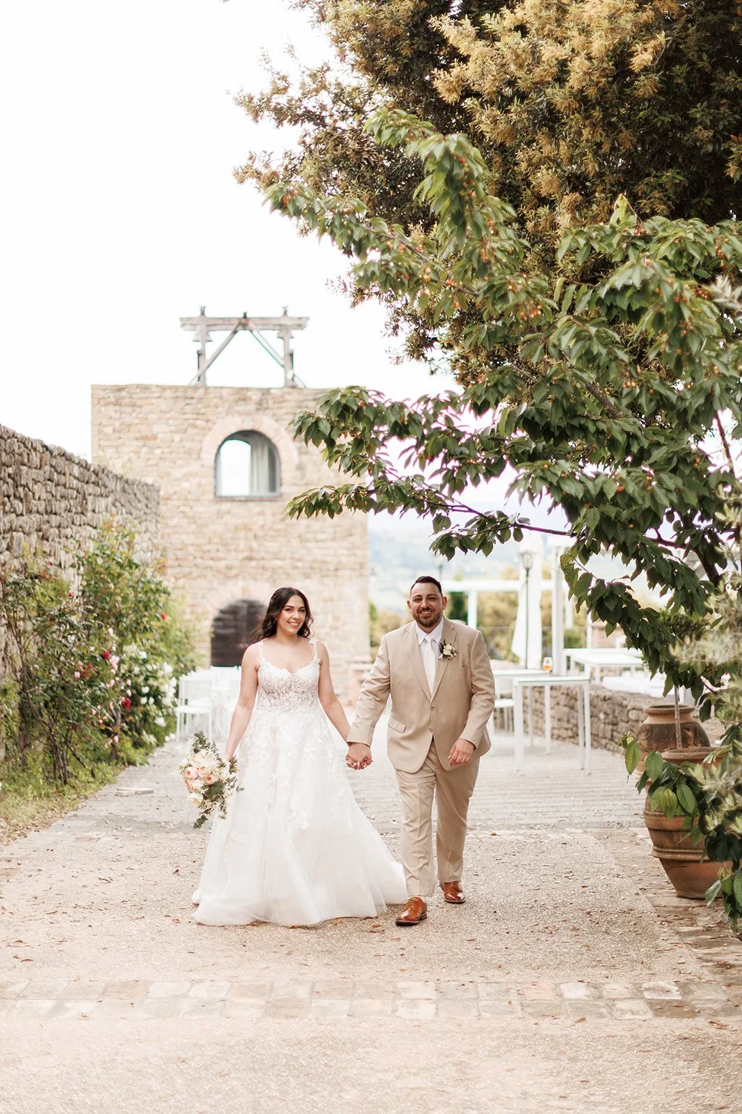 A bride and groom holding hands and smiling while walking outside during their wedding. The bride wears a white lace wedding dress and holds a bouquet of flowers. The groom wears a beige suit with a matching tie and brown shoes. There are trees, ston