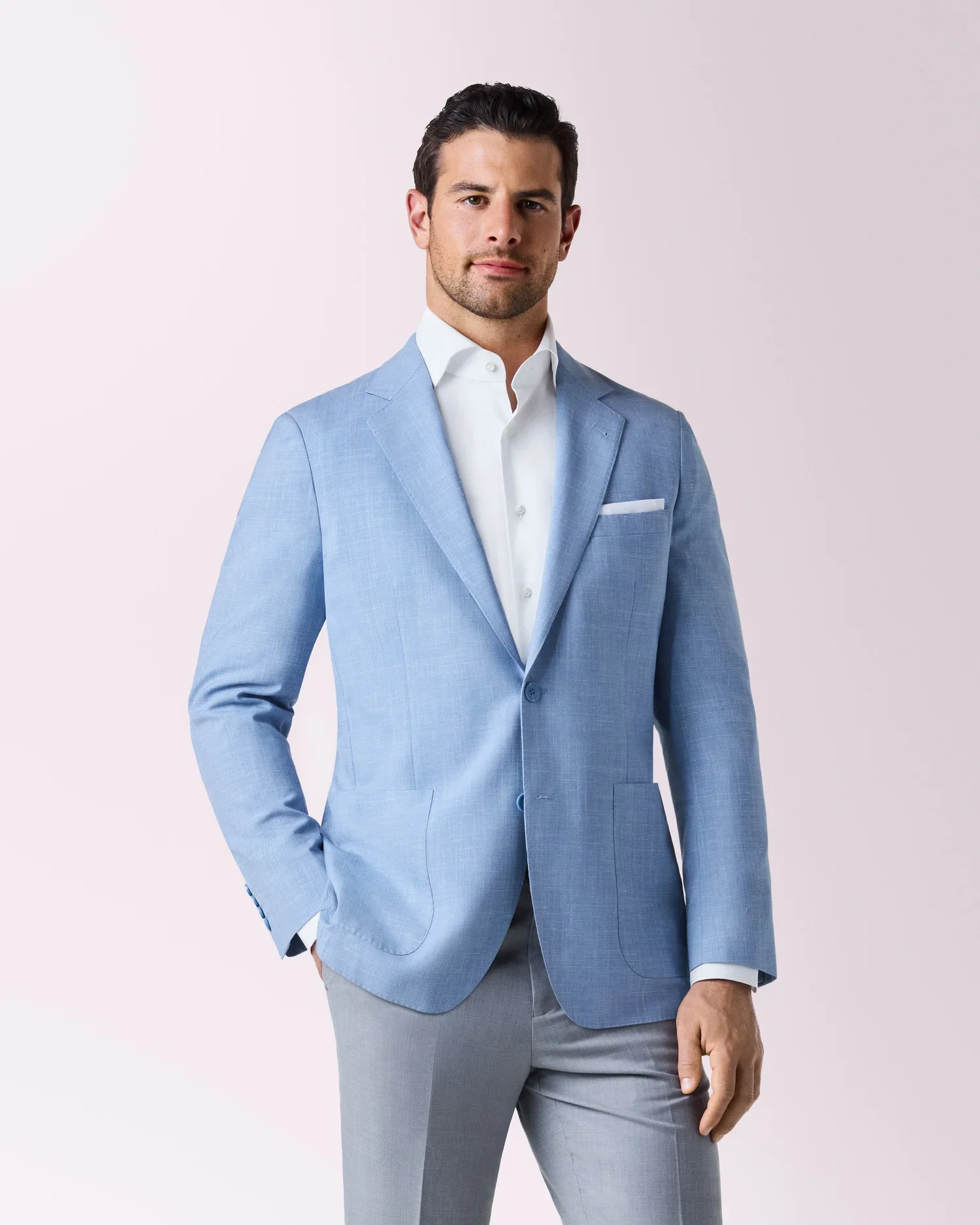 A man in a light blue blazer, white shirt, and gray pants standing against a plain light background.