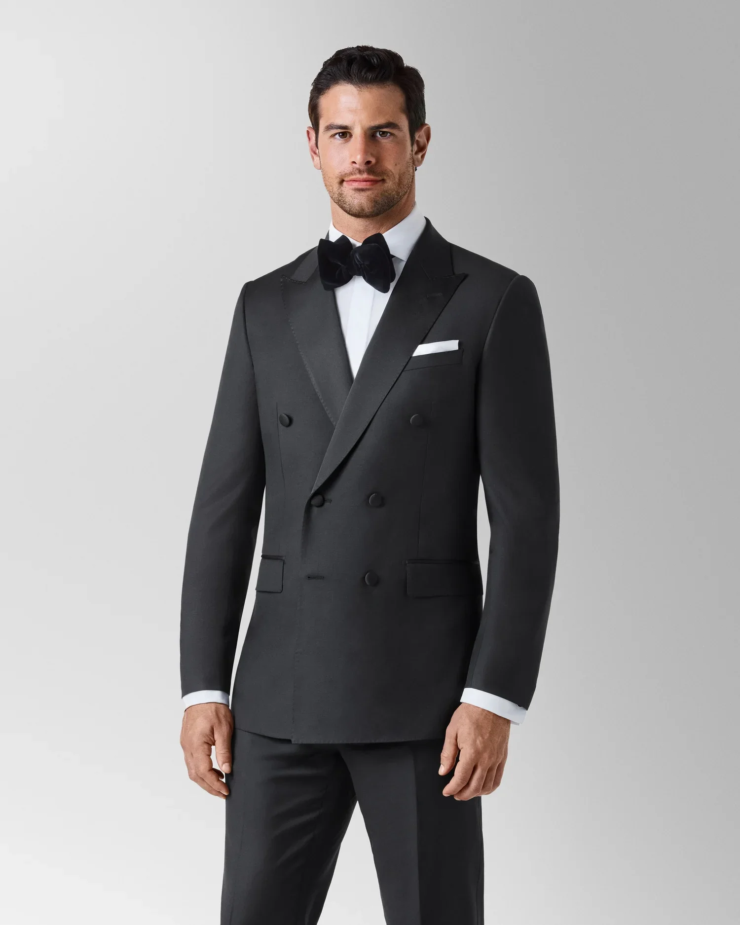 A man wearing a black tuxedo with a white dress shirt, black bow tie, and pocket square, standing against a light gray background.