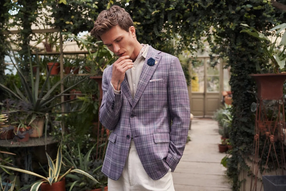 Young man in a checkered blazer with a flower lapel pin, standing in a greenhouse with plants and potted greenery.