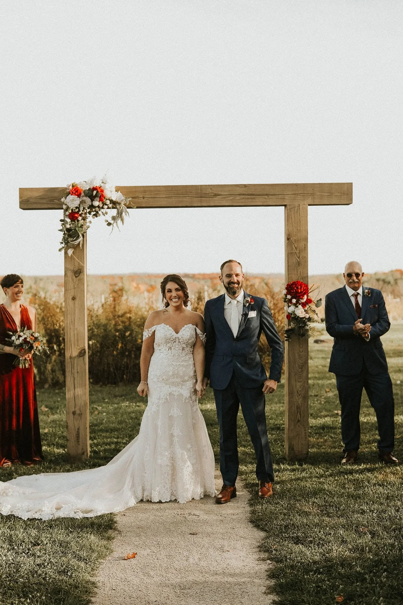 A bride and groom holding hands and smiling at an outdoor wedding ceremony with a wooden arch, decorated with flowers, in the background.