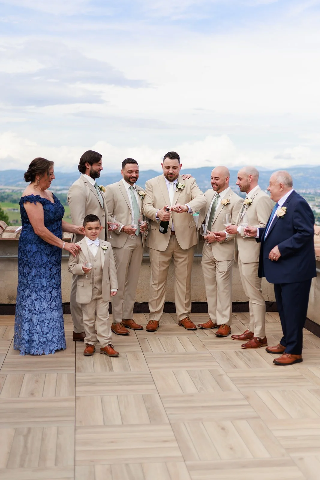 A wedding celebration with the groom, groomsmen, mother, and a young ring bearer standing on a balcony with scenic mountain views, some holding glasses and the groom opening a bottle of champagne.