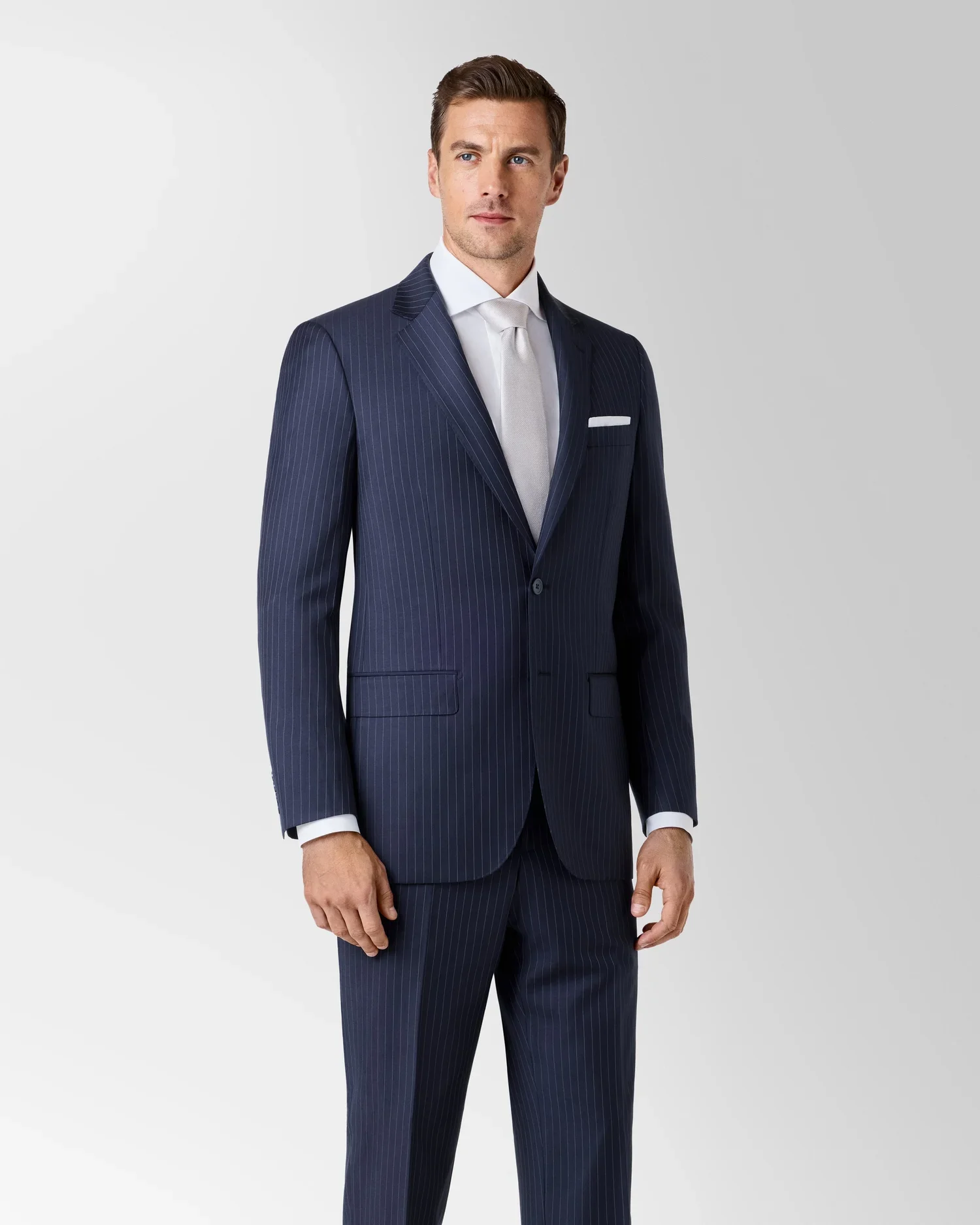 Man in a navy blue pinstripe suit with a white shirt and white tie, standing against a plain light gray background.