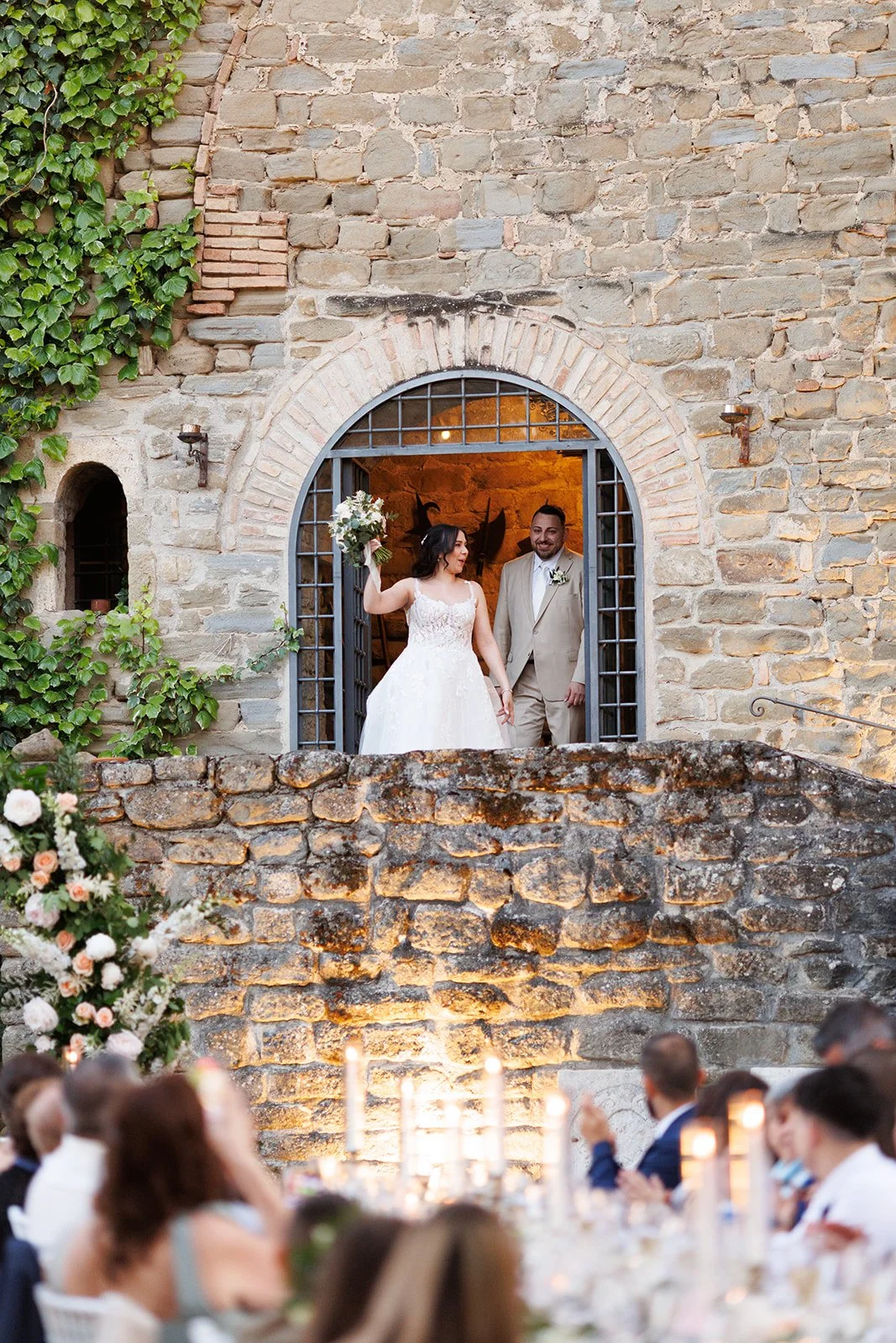 A bride and groom standing in an arched doorway during their wedding reception. The bride is holding a bouquet and smiling while the groom looks at her. Guests are seated at tables below, decorated with candles and flowers.