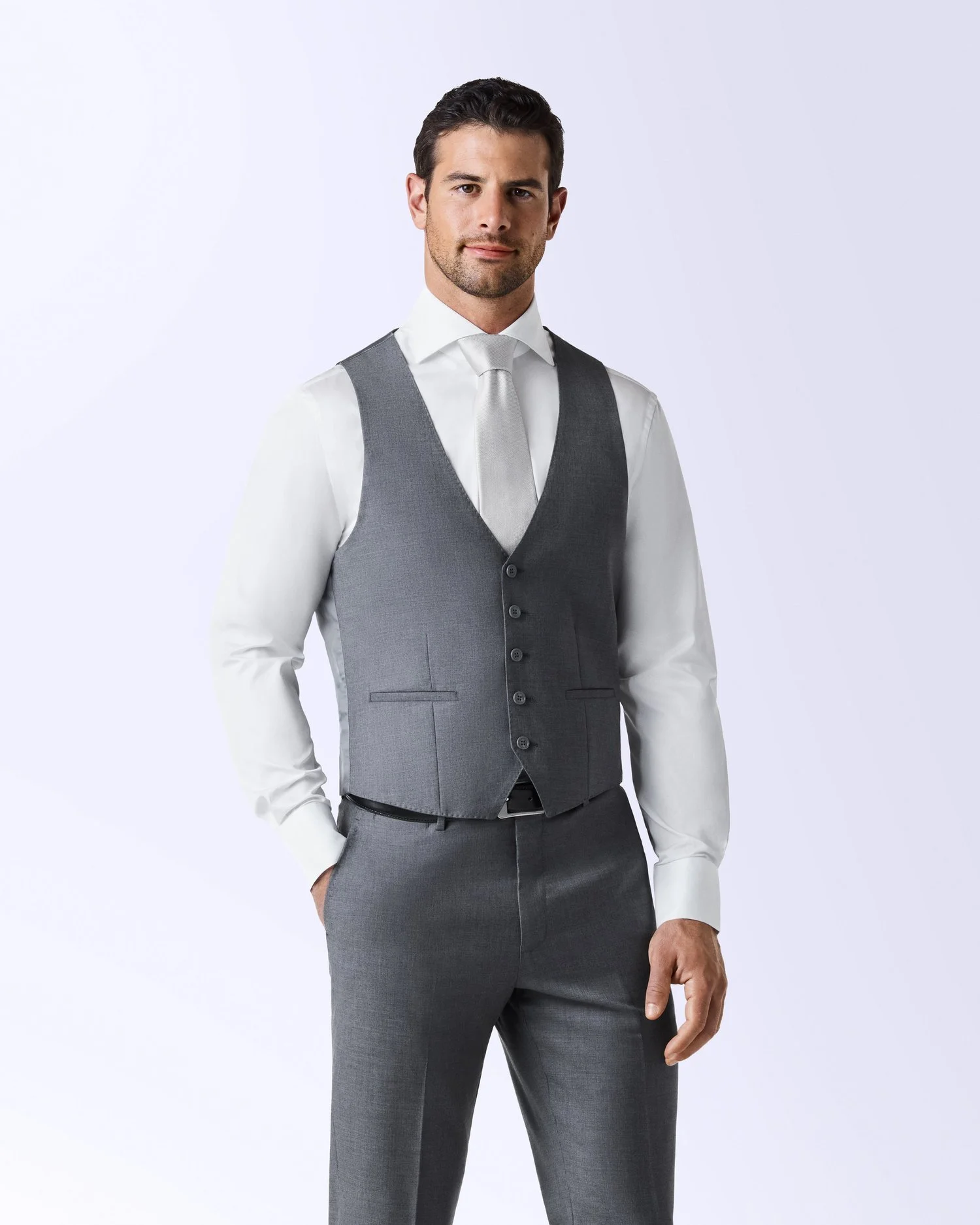 A man dressed in a white shirt, silver tie, and gray vest and pants, standing against a plain light background.