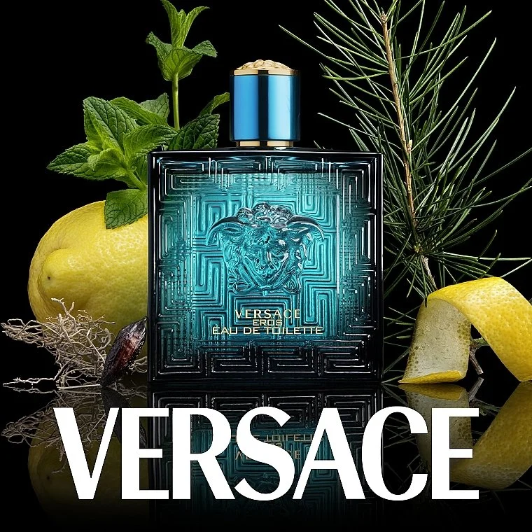 A bottle of Versace Eros Eau de Toilette perfume with citrus fruits and green foliage around it on a reflective surface.