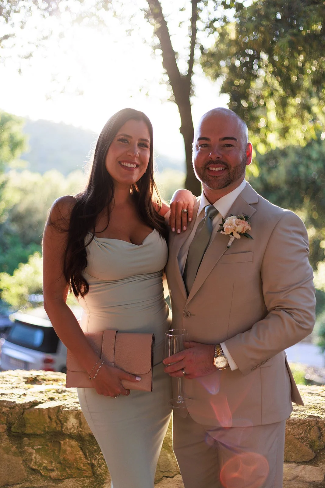 A happy couple dressed in formal attire outdoors during daytime, with the woman wearing a strapless light-colored dress holding beige clutch and the man in a beige suit with a boutonniere, holding a glass of champagne.