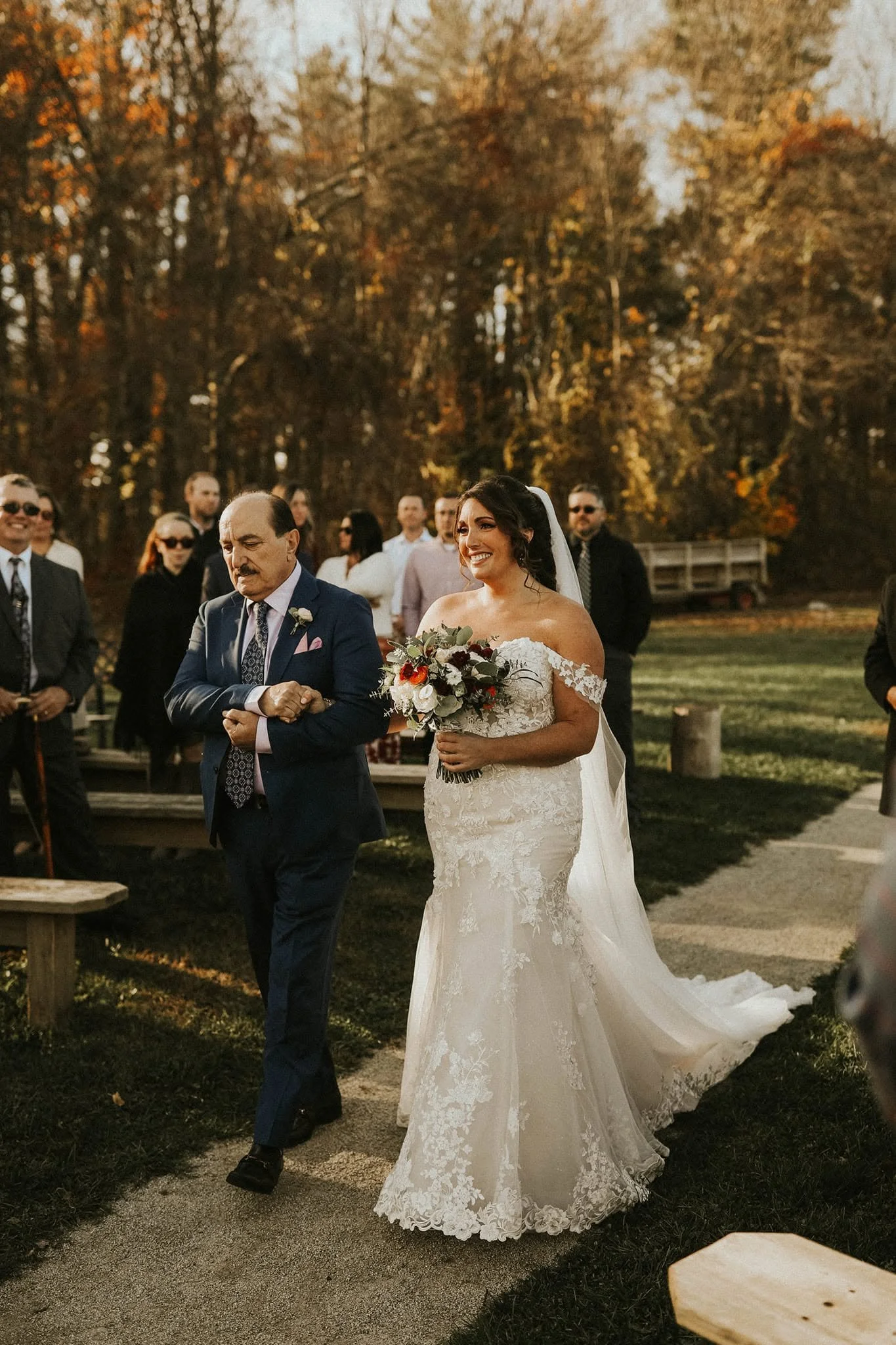 A bride in a white lace wedding dress holding a bouquet, walking outdoors with a man in a dark suit, during a wedding ceremony with guests in the background and autumn trees.
