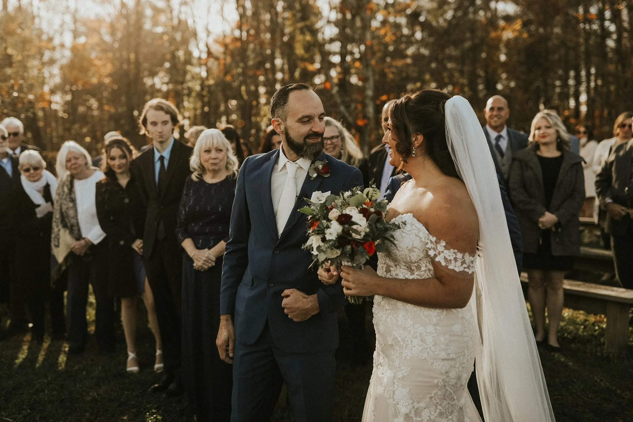 A bride and groom stand close together outdoors, holding hands, during their wedding ceremony as family and friends look on, with autumn trees in the background.