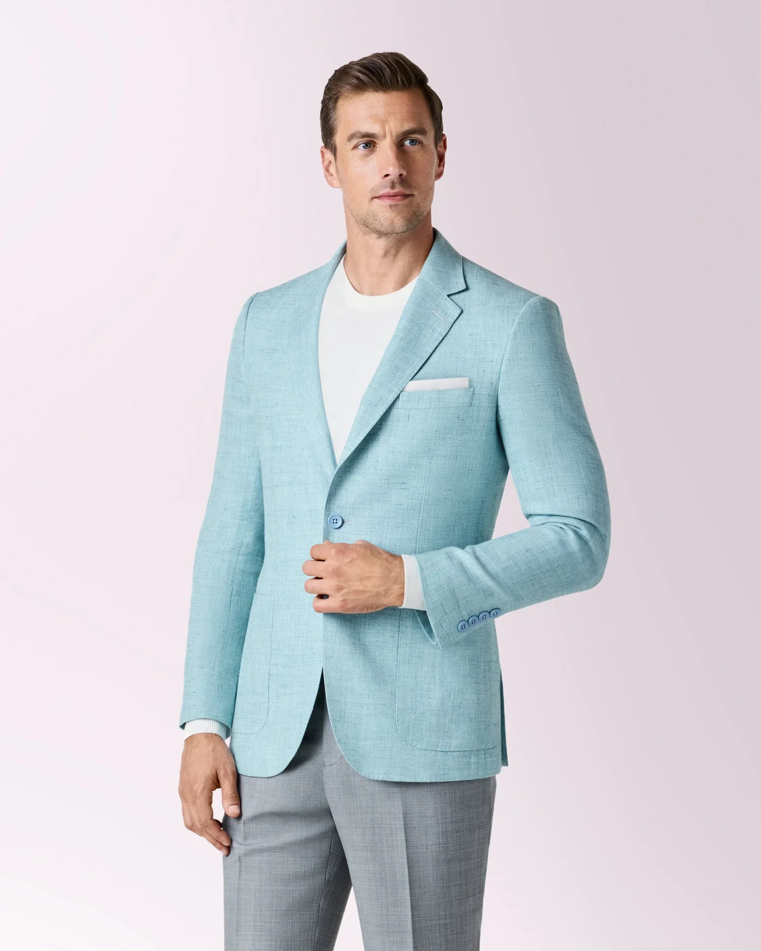 A man dressed in a light blue blazer, white shirt, and gray pants standing against a plain light-colored background.