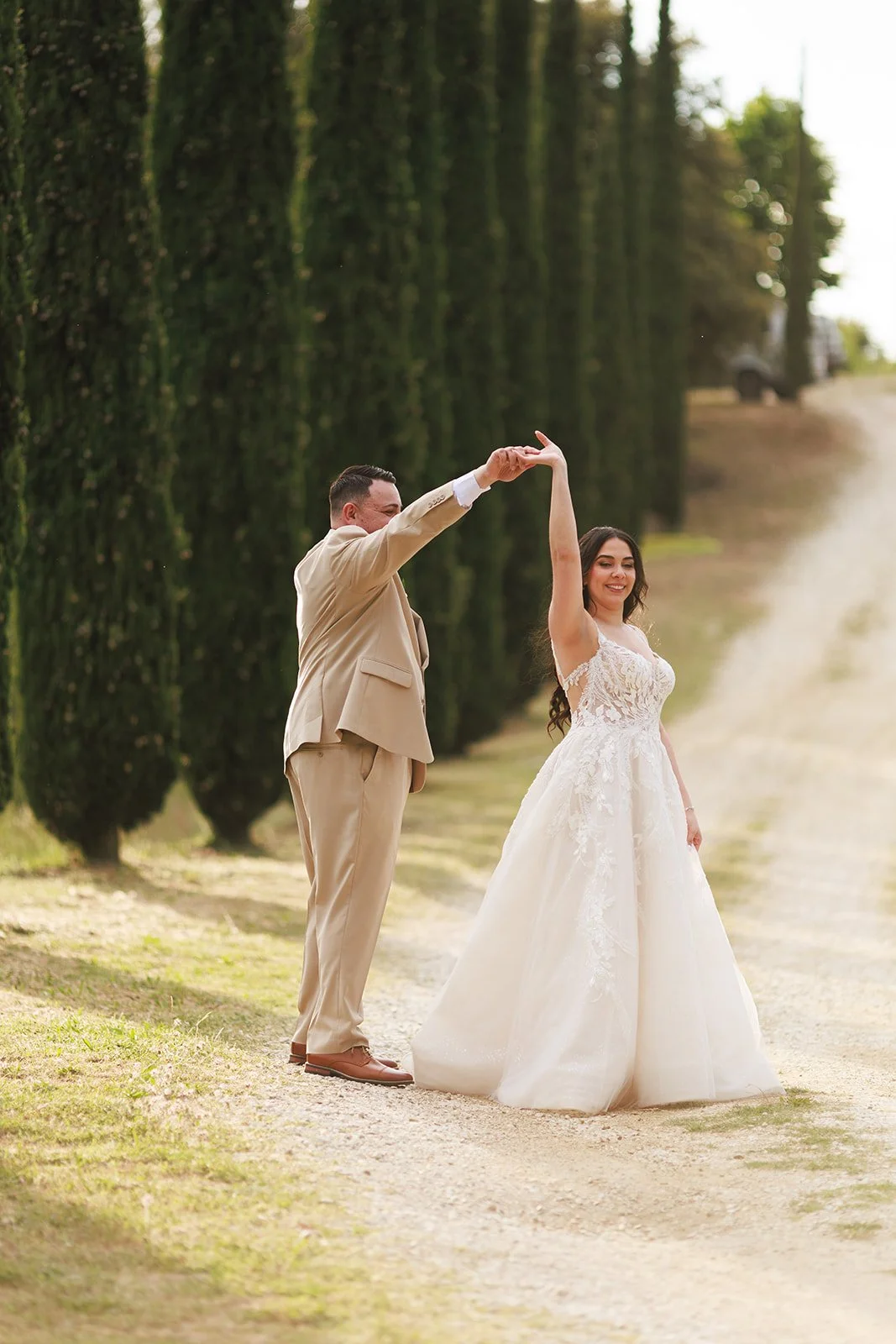 A newlywed couple dancing outdoors on a dirt path lined with tall cypress trees, with the groom in a beige suit and the bride in a white lace wedding gown.