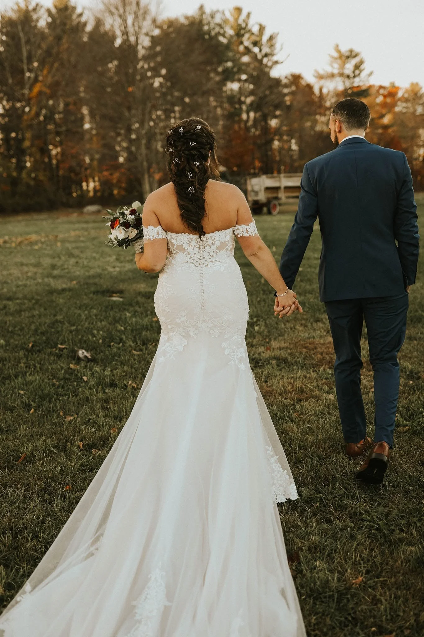A bride and groom walking hand in hand outdoors during sunset, with the bride wearing an off-shoulder lace wedding gown and holding a bouquet, and the groom in a navy suit. The background shows trees with autumn foliage and a trailer.