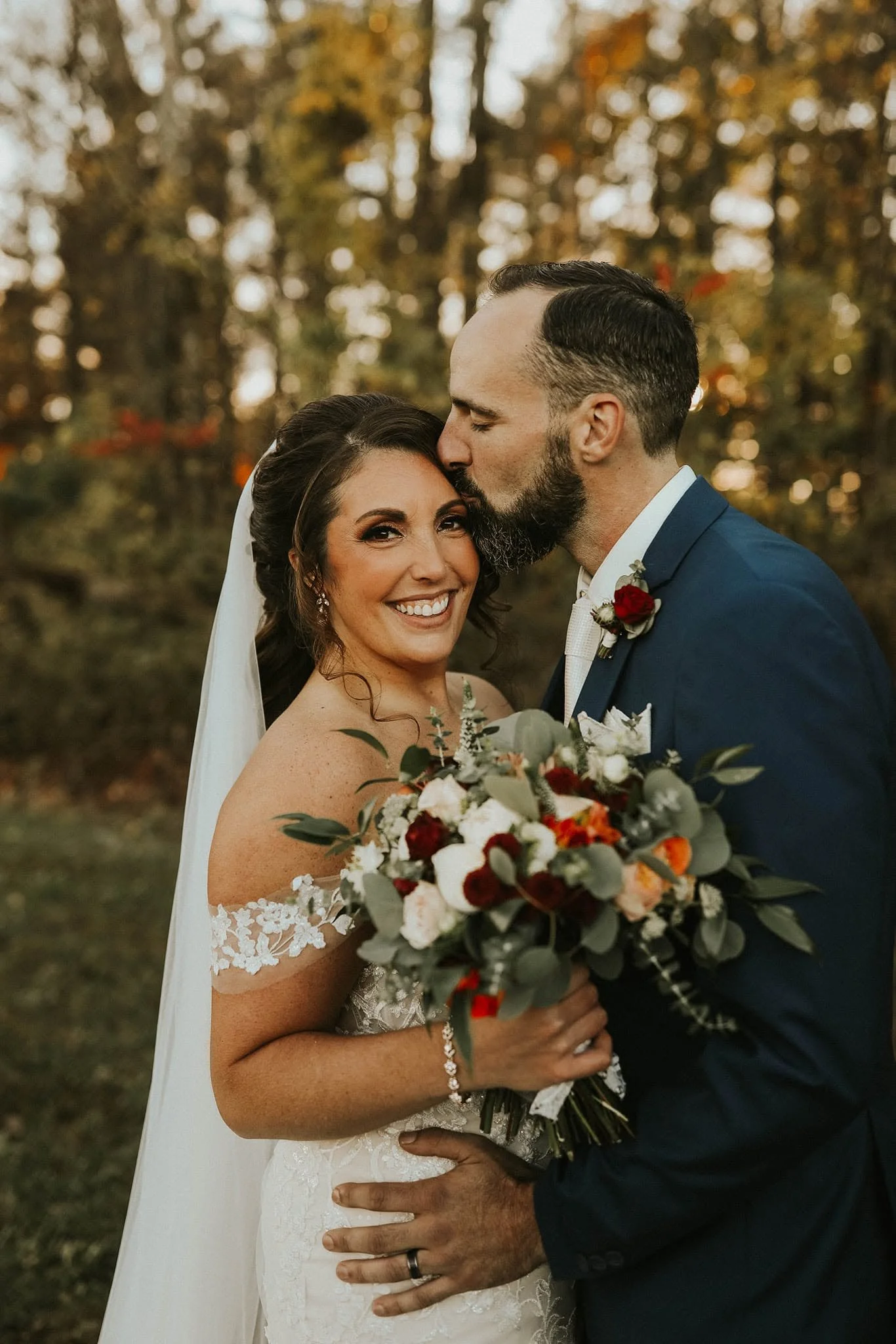 A bride and groom are standing outdoors in front of a blurred background of autumn trees. The groom is kissing the bride on her forehead. The bride is smiling and holding a large bouquet of flowers in front of her. The bride is wearing a white lace w