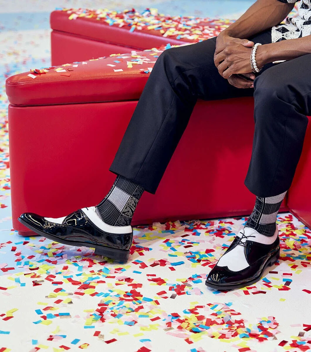 A person sitting on a red bench with confetti scattered on the floor and bench. They are wearing black pants, black and white dress shoes, patterned socks, a watch on their wrist, and a bracelet.