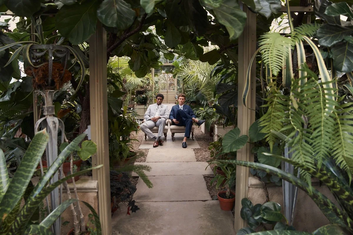 Two men sitting on a sofa inside a greenhouse surrounded by lush green plants and ferns.