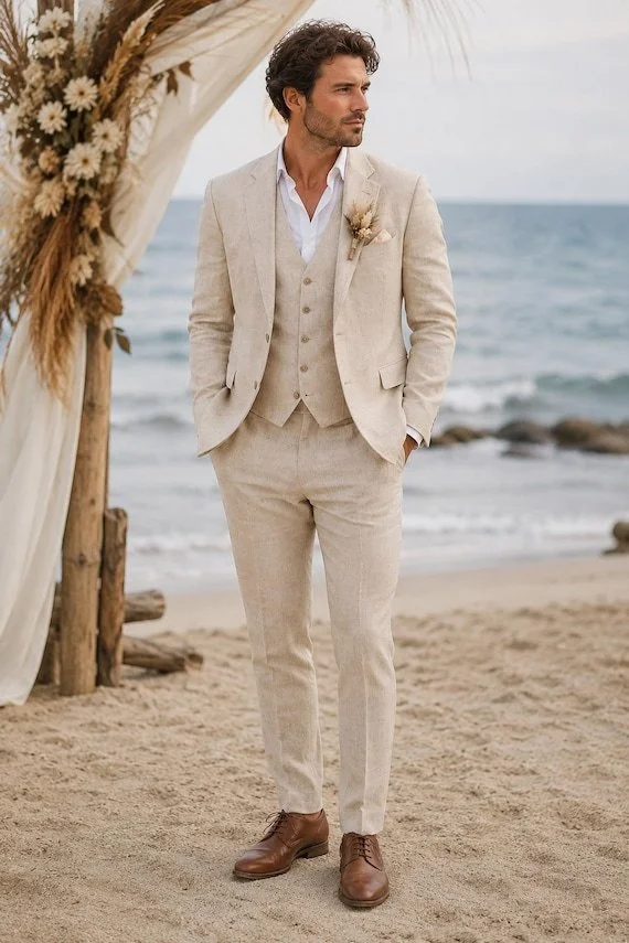 A man dressed in a light-colored suit standing on a sandy beach, with the ocean in the background, under a decorated arch with flowers and dried plants.