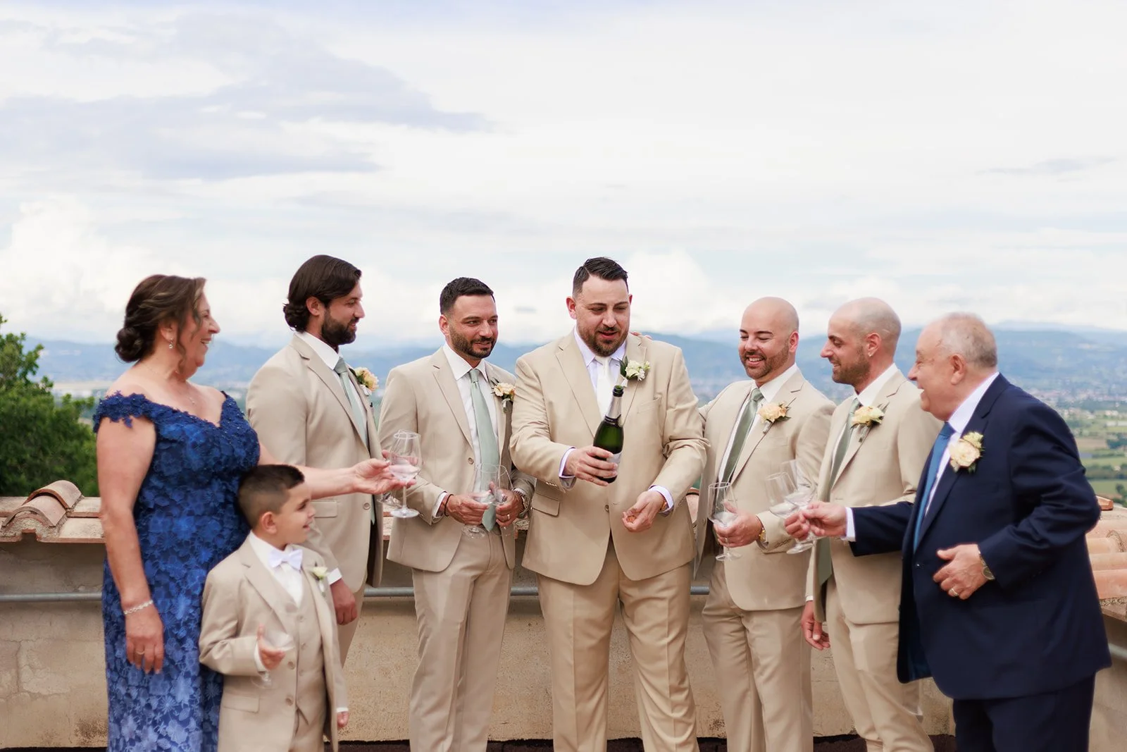 A group of people at a wedding celebration, including women and men in suits, standing outdoors on a balcony with a scenic view of the mountains and landscape in the background, toasting with glasses and smiling.