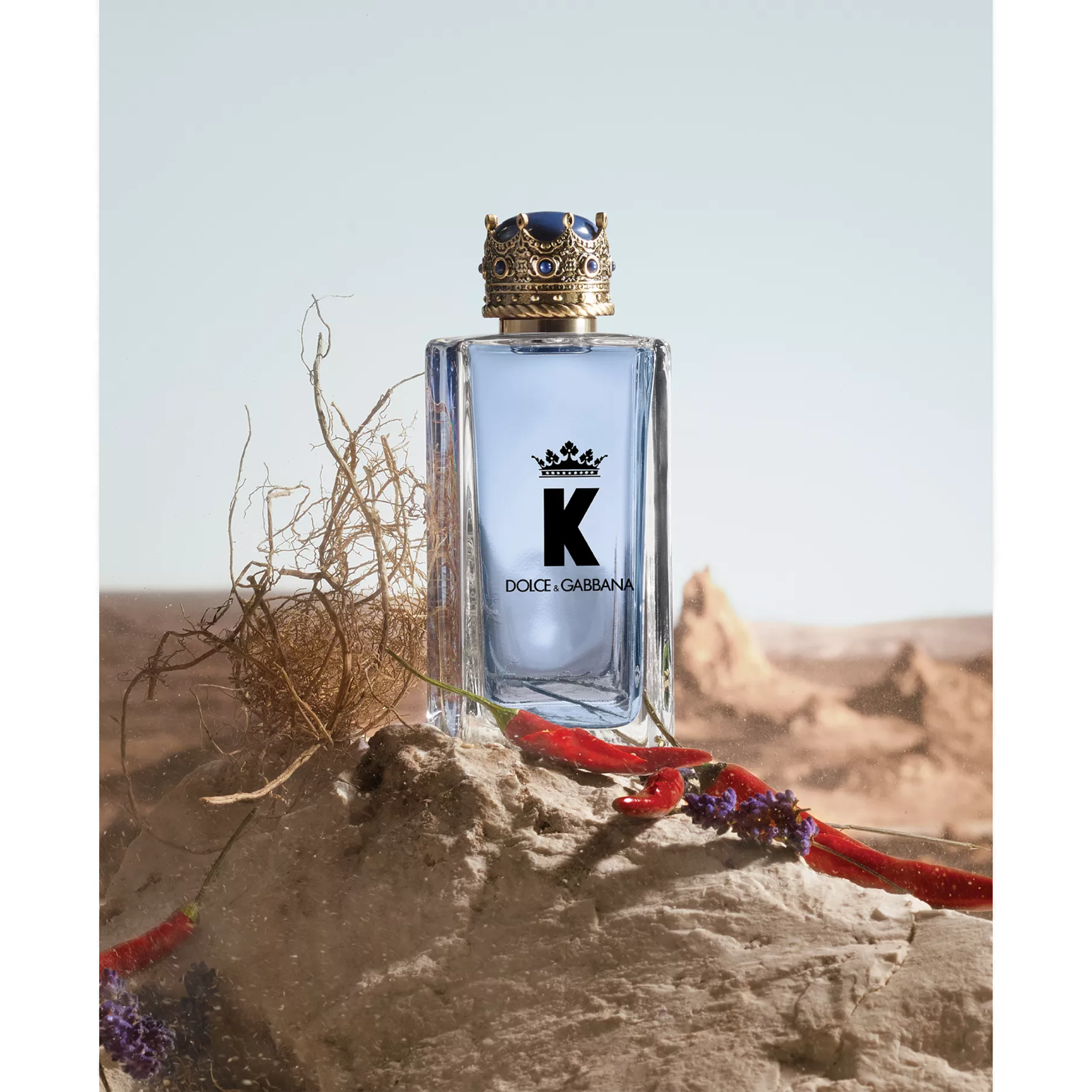 A clear glass perfume bottle with a decorative gold crown-like cap, set against a neutral background with earthy elements like roots and red peppers.