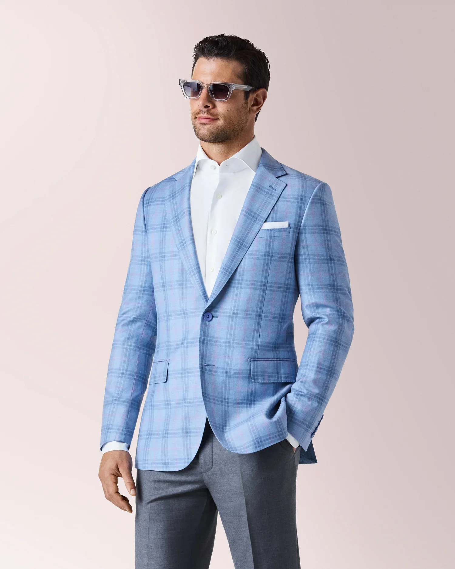 A man wearing a light blue plaid suit jacket, white dress shirt, gray dress pants, and sunglasses poses against a plain background.