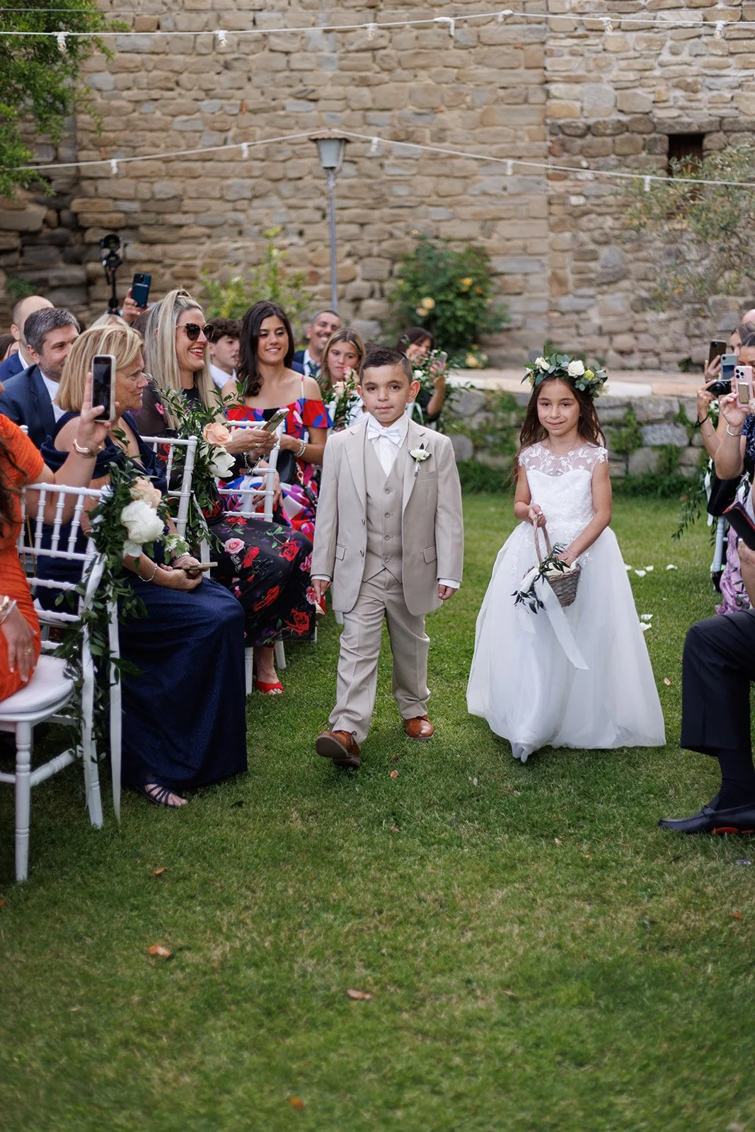 Two children, a boy in a beige suit and a girl in a white dress with a floral crown, walking down the aisle at a wedding ceremony, surrounded by seated guests taking photos.