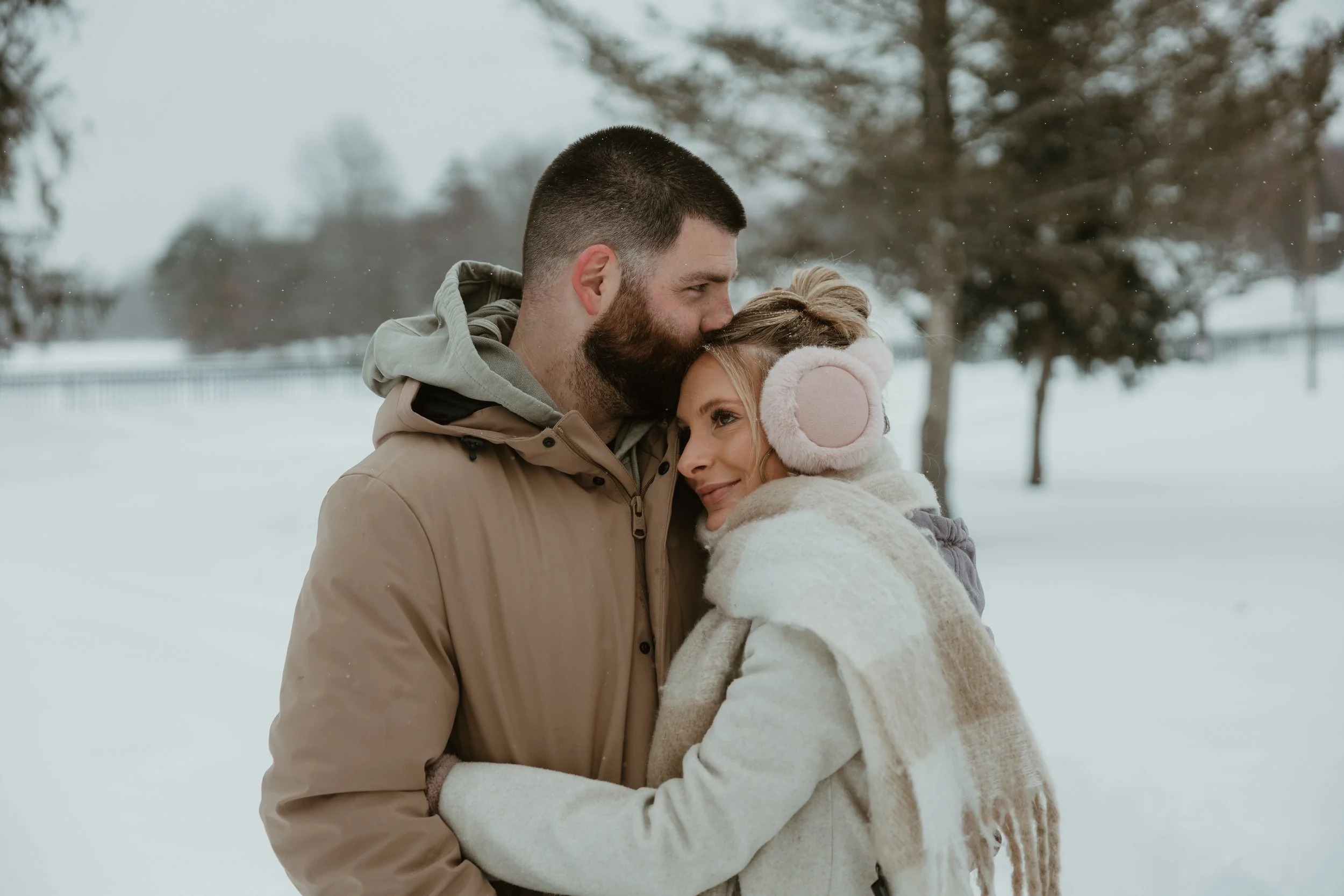 A man and woman embrace outdoors in a snowy park, with trees and a fence in the background.