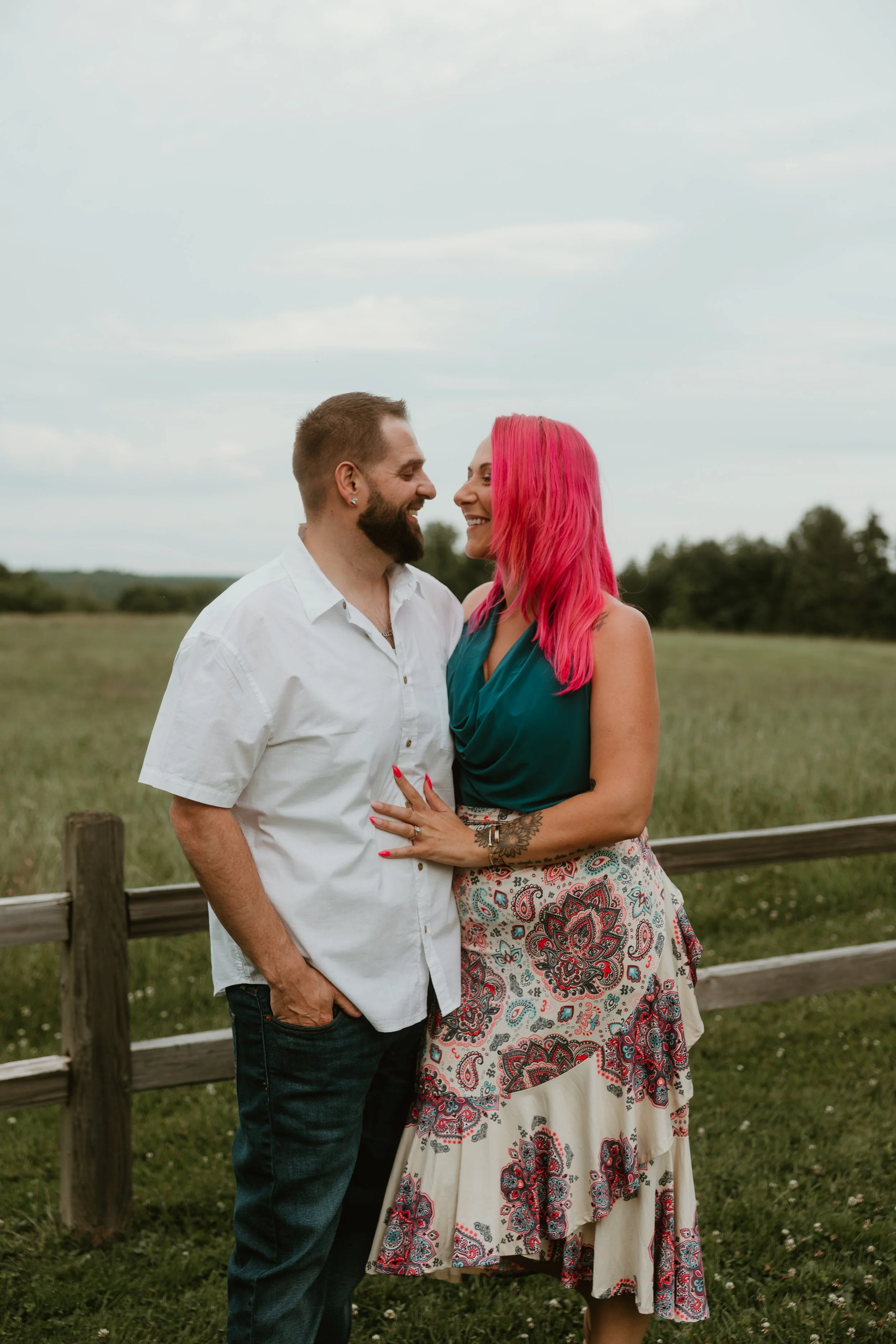 A couple smiling and gazing at each other outdoors in a grassy field with a wooden fence and trees in the background