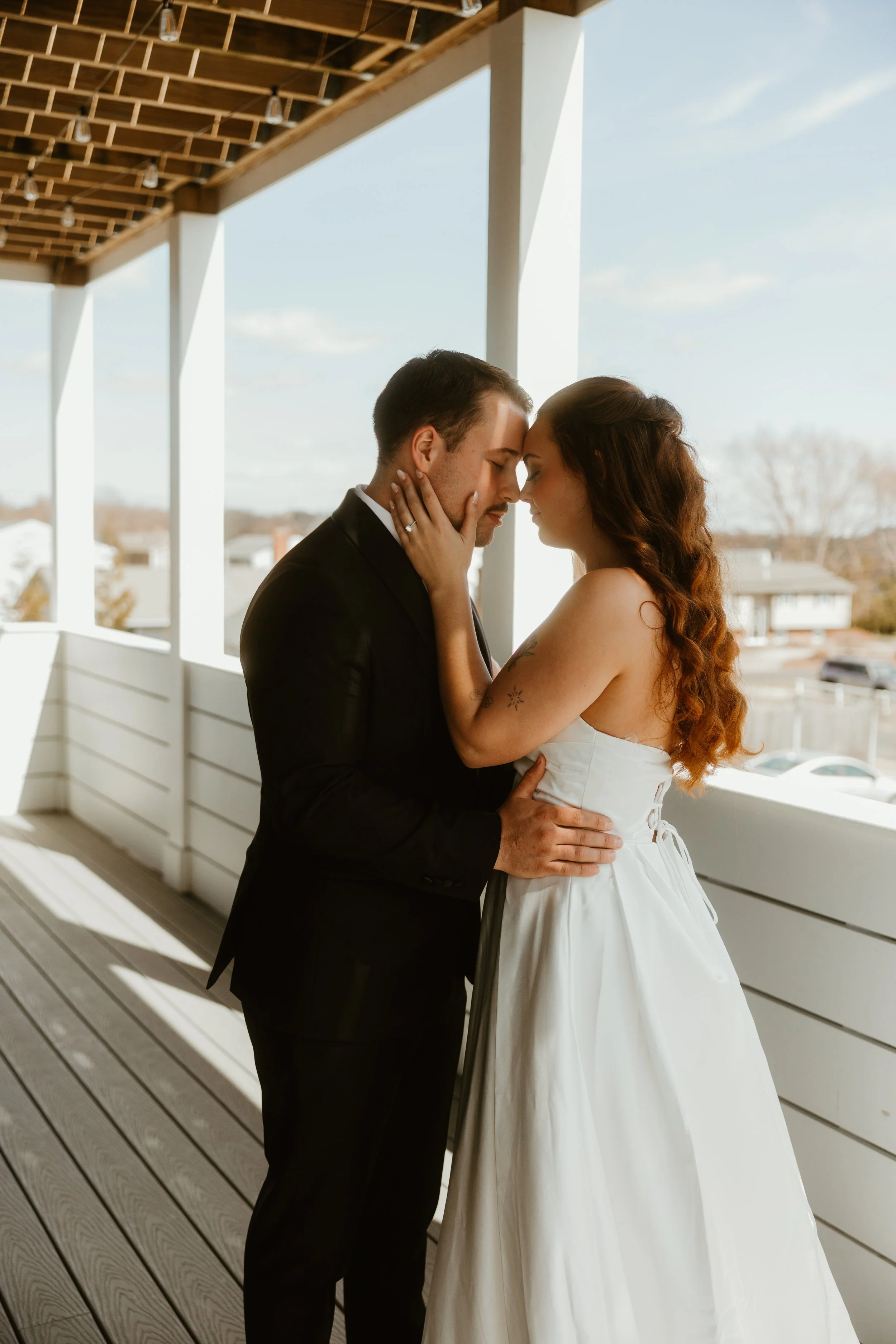 A bride and groom share a close, intimate moment on a balcony with a white wooden railing and a roof with string lights, overlooking a suburban neighborhood during daytime.