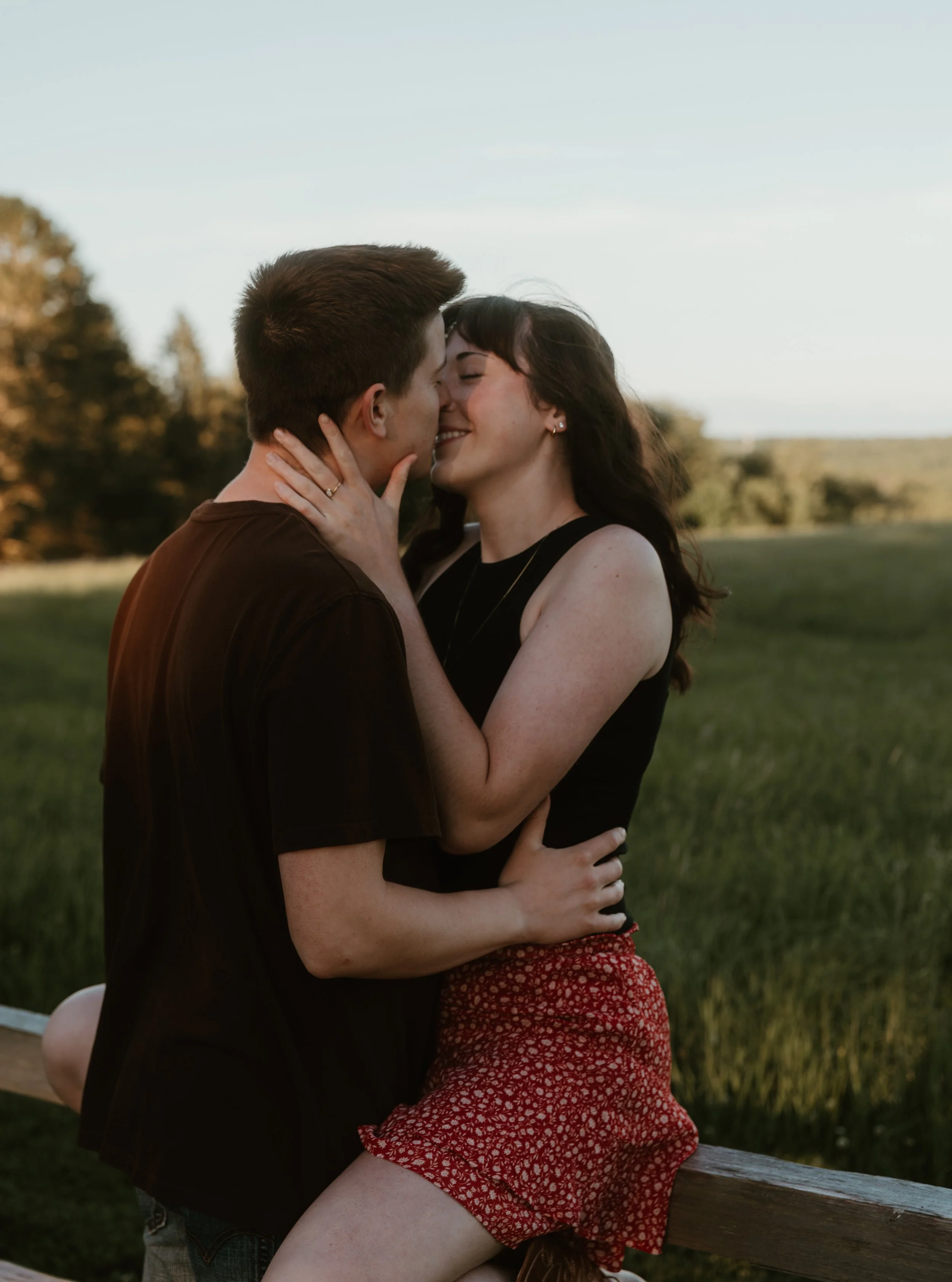 A young couple is kissing outdoors in a park or field during sunset, with trees and grass in the background.