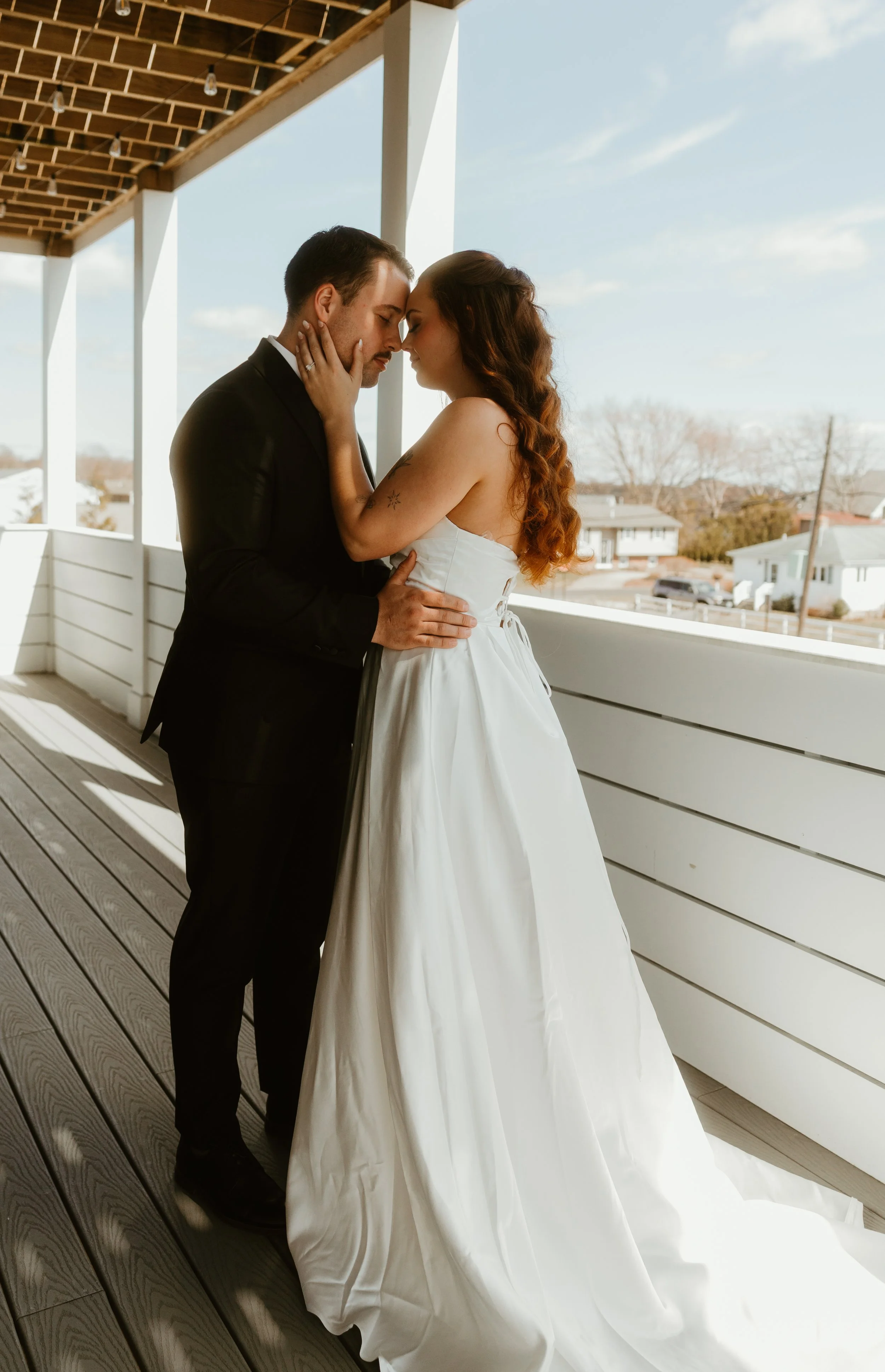 A bride and groom with foreheads touching, eyes closed, on a balcony during daytime. The bride is wearing a strapless white wedding gown with a full skirt; the groom is in a black suit and tie. The bride has long, curly hair and a tattoo on her upper