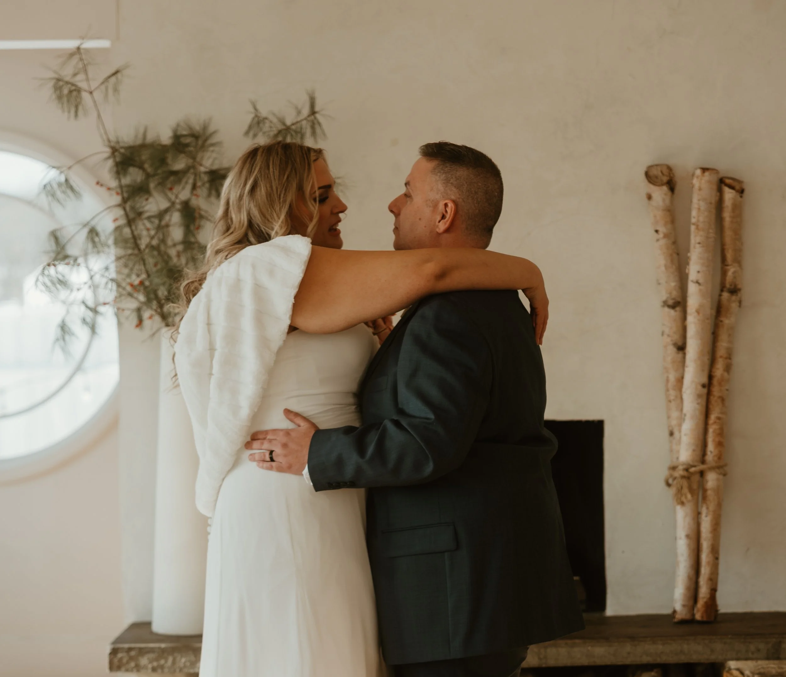 A woman in a white dress embraces a man in a dark suit indoors, with a neutral wall, a window, and decorative branches in the background.