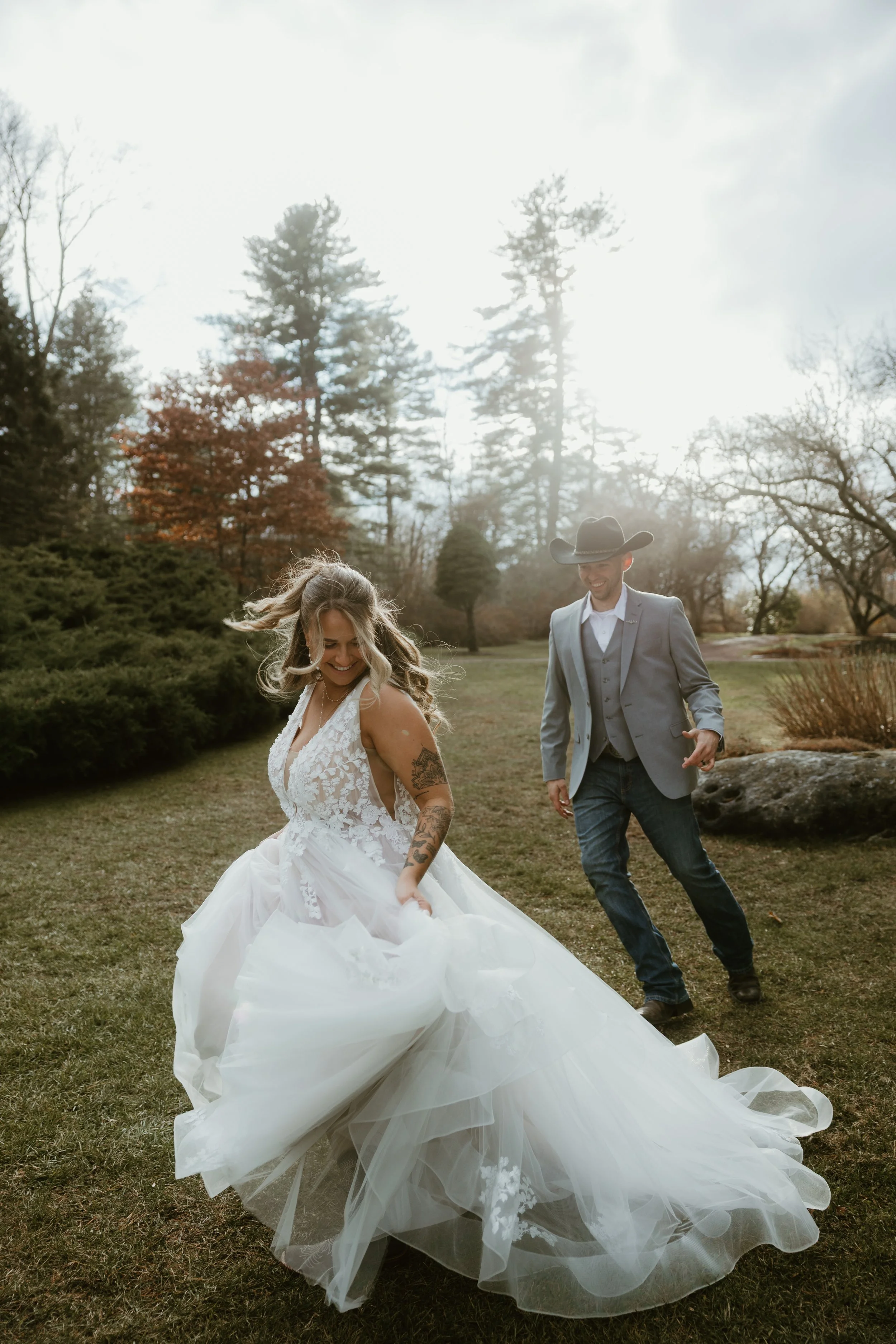 A bride in a white wedding dress joyfully spins in a garden with green grass, trees, and autumn-colored foliage, while a man in a gray suit and cowboy hat smiles nearby.