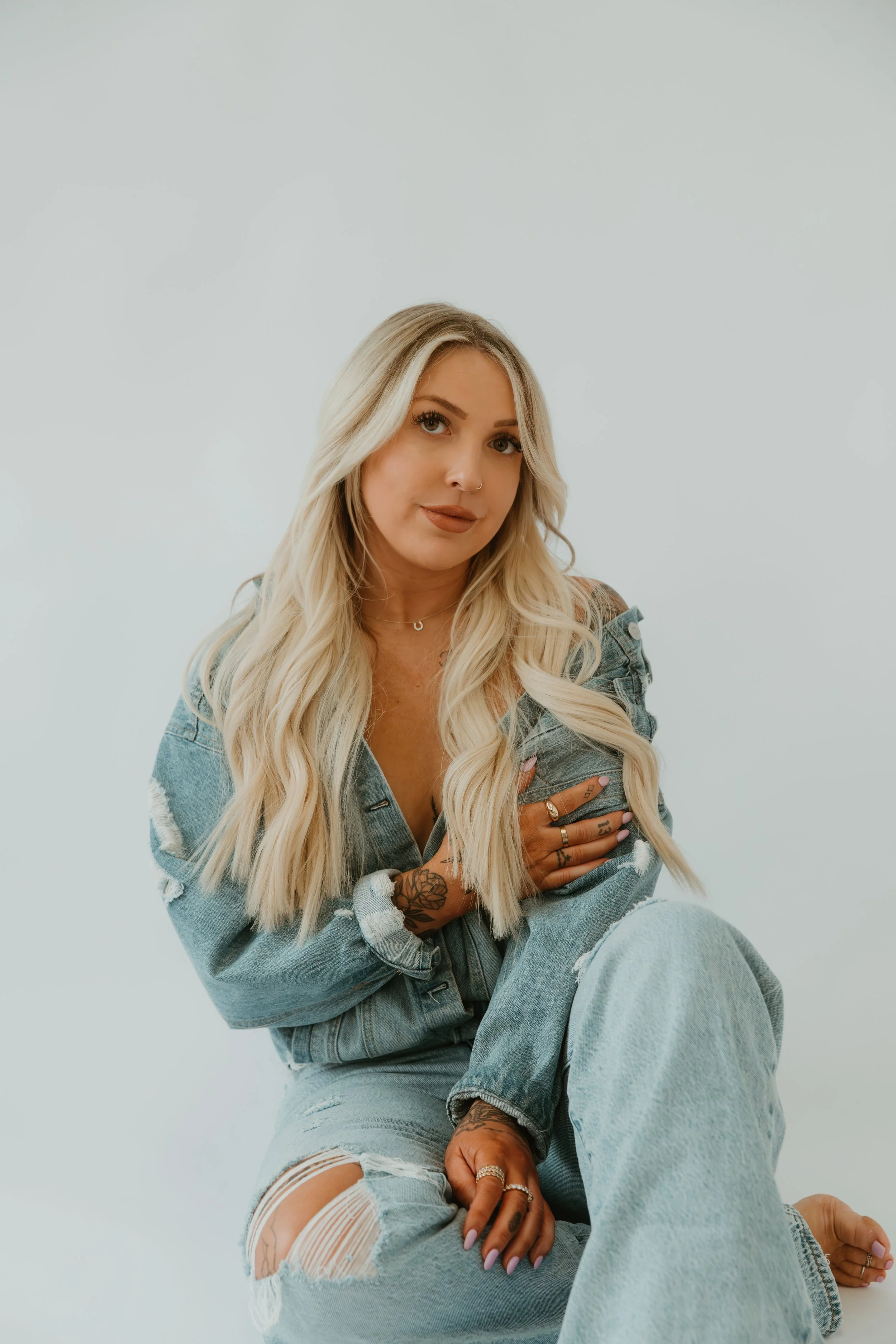 A young woman with long blonde wavy hair, wearing a distressed denim jacket and ripped jeans, sitting against a plain light-colored background.