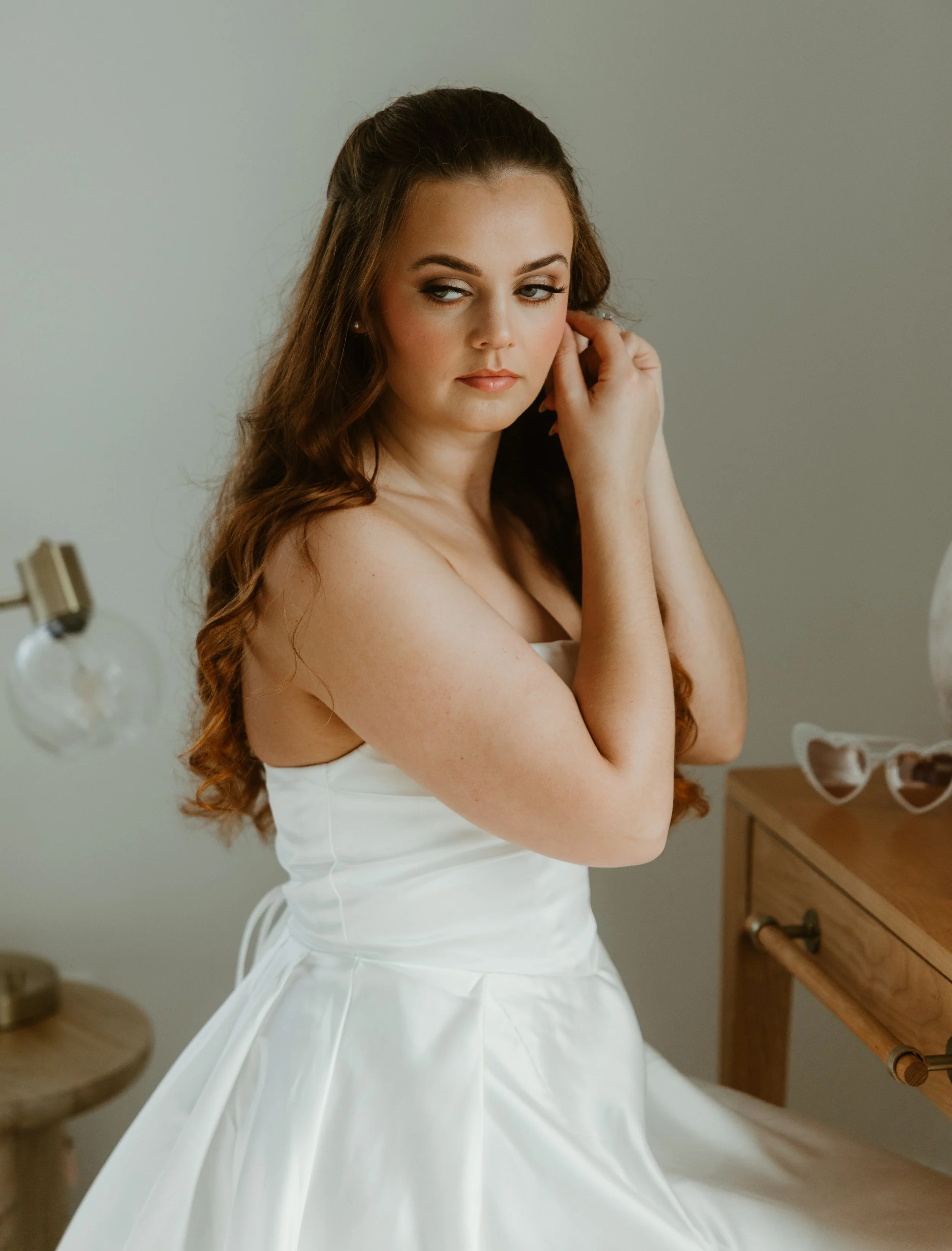 A young woman with long, curly red hair, wearing a white dress, gazes softly to the side and touches her ear in a softly lit room.