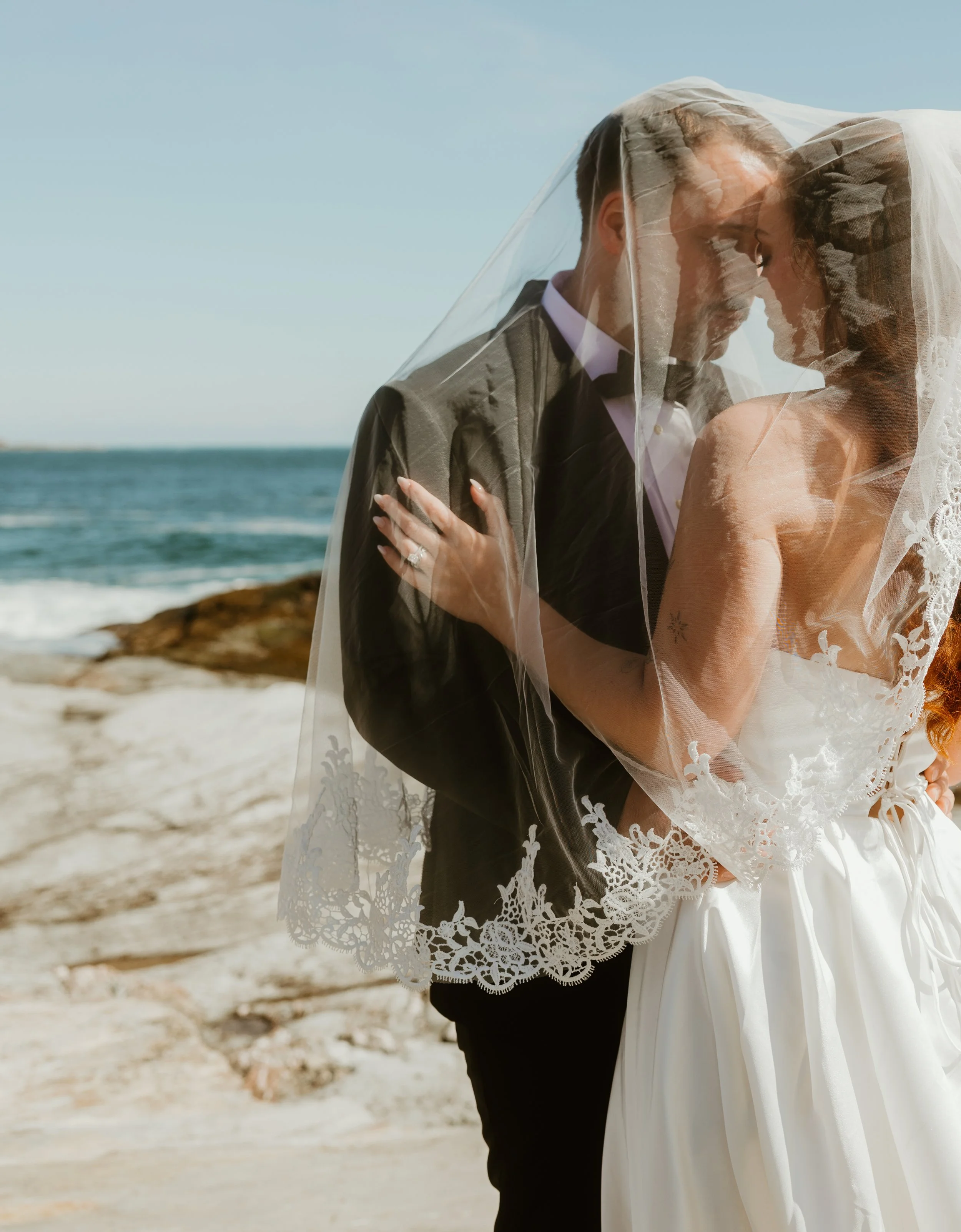 A bride and groom kissing under a veil on a beach with sand and ocean in the background.
