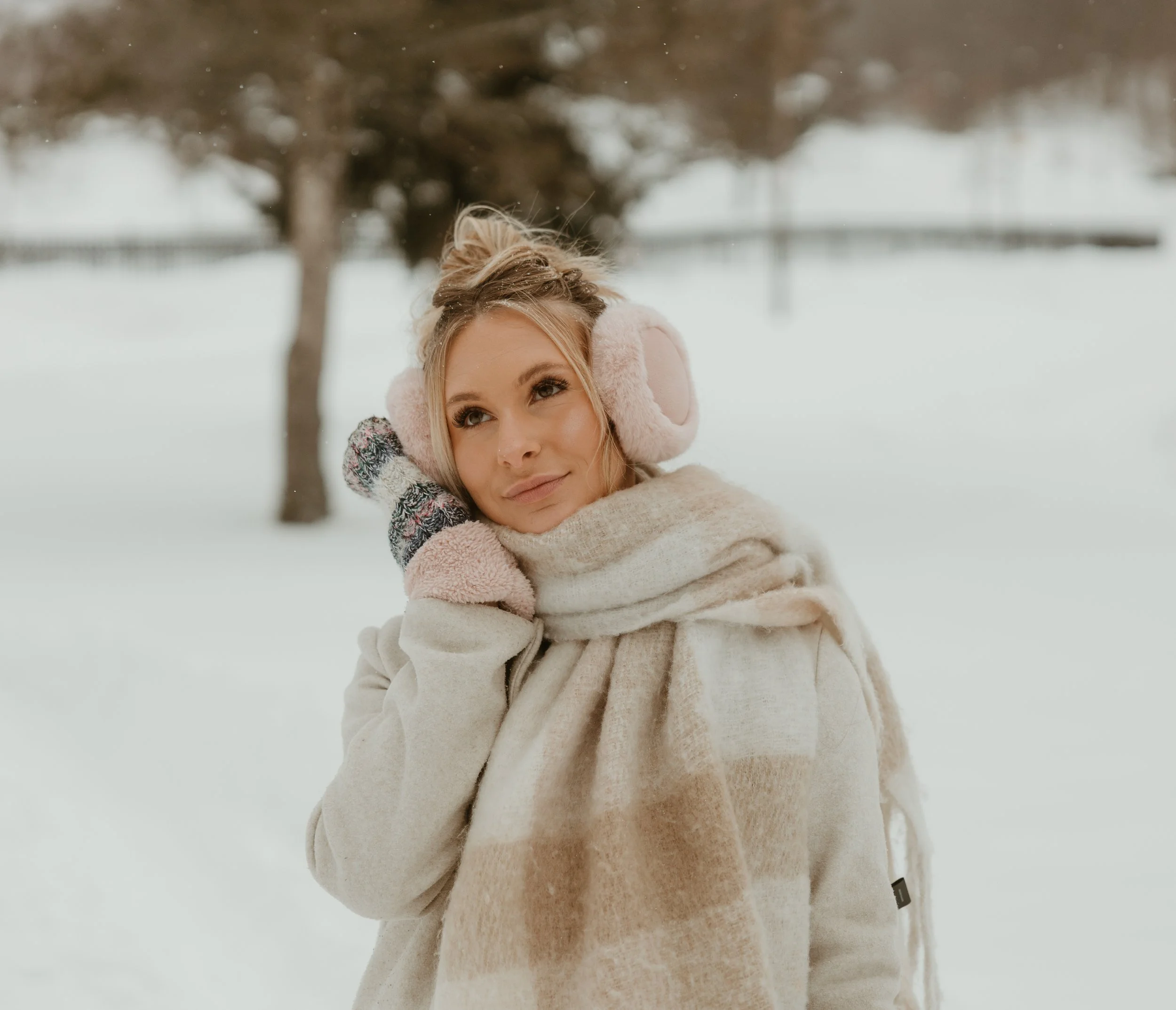 A woman with blonde hair wearing pink earmuffs and winter coat outdoors in a snowy landscape with blurred trees in the background.