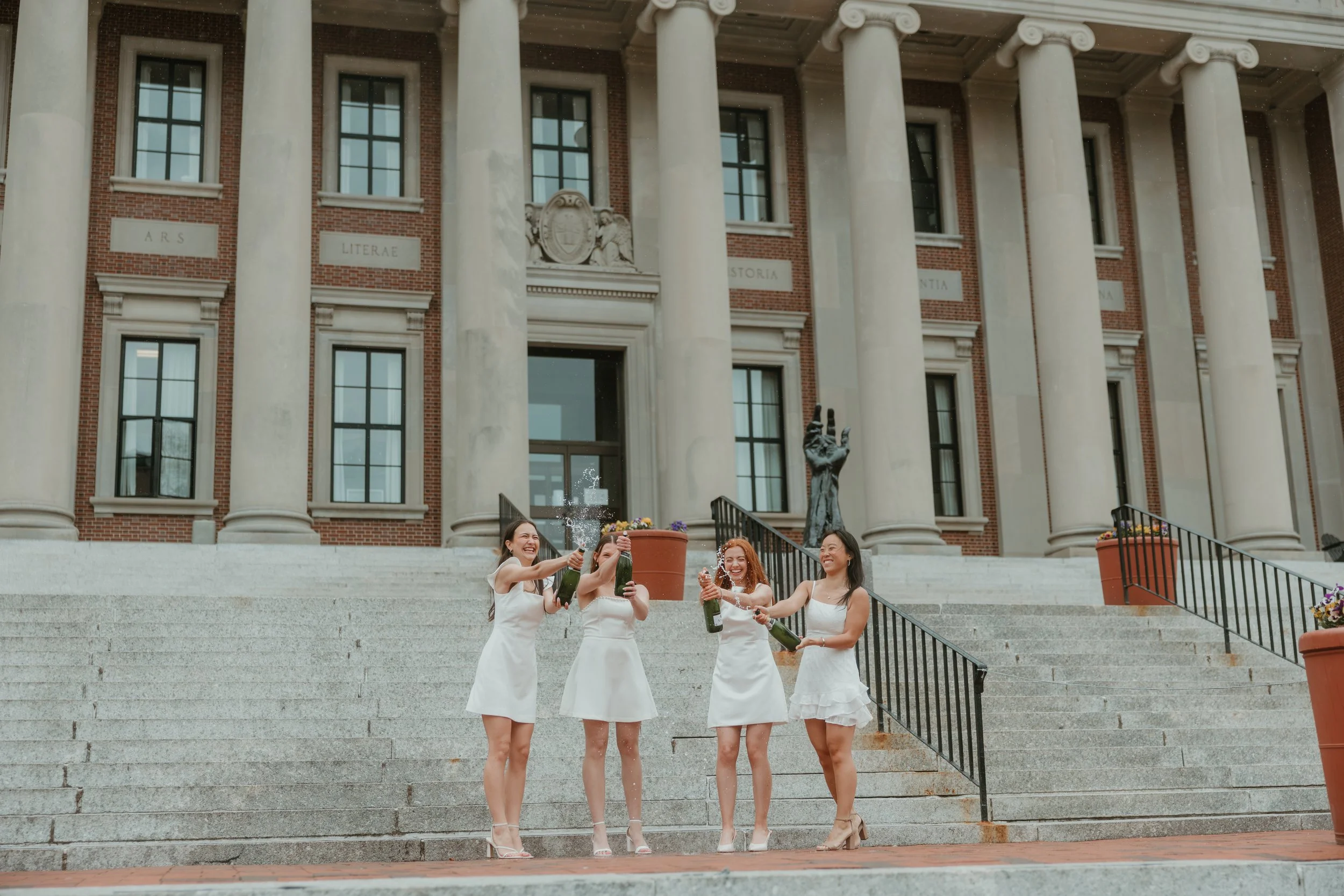 Four women in white dresses celebrating and toasting with bottles of champagne on the steps of a historic building with large columns and an inscription reading 'ARS LITerae' and 'HISTORIA' above the entrance.