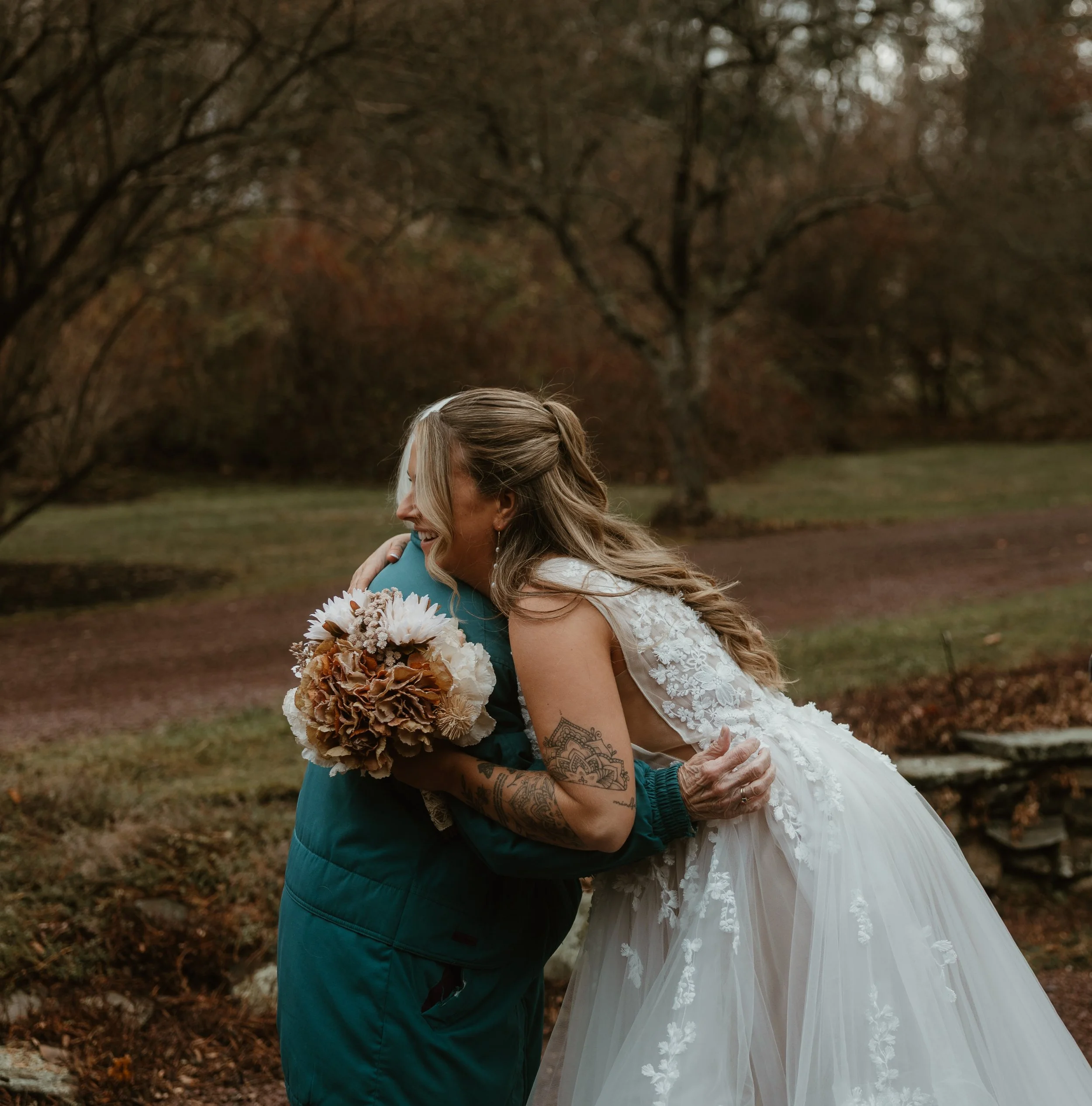 A groom and bride embrace outdoors during a wedding, with the groom in a dark jacket and the bride in a white lace wedding dress holding a bouquet of flowers, against a background of autumn trees.