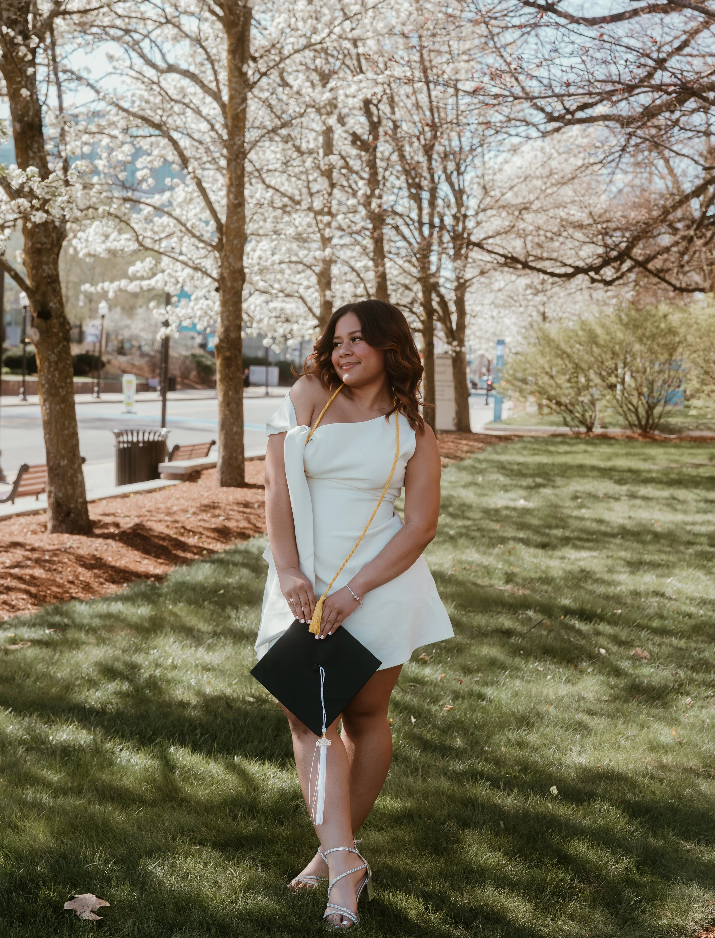 A woman in a white dress and heels holding a graduation cap, standing on grass, with blooming trees and a sidewalk in the background.