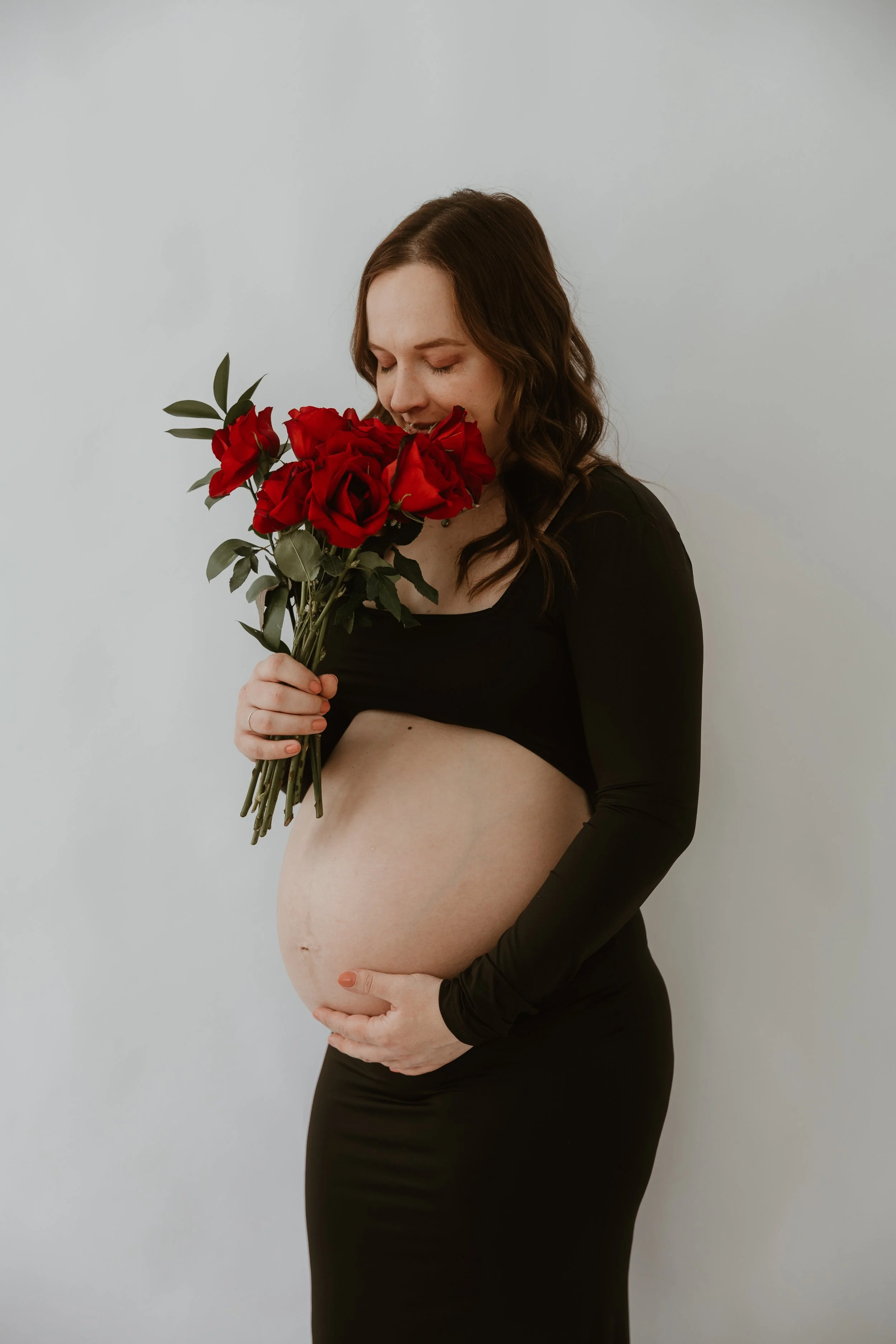 A pregnant woman with wavy brown hair holding a bouquet of red roses and a few green leaves. She is wearing a black long-sleeve top and black pants, standing against a plain light gray background. She is gently touching her baby bump with her right h