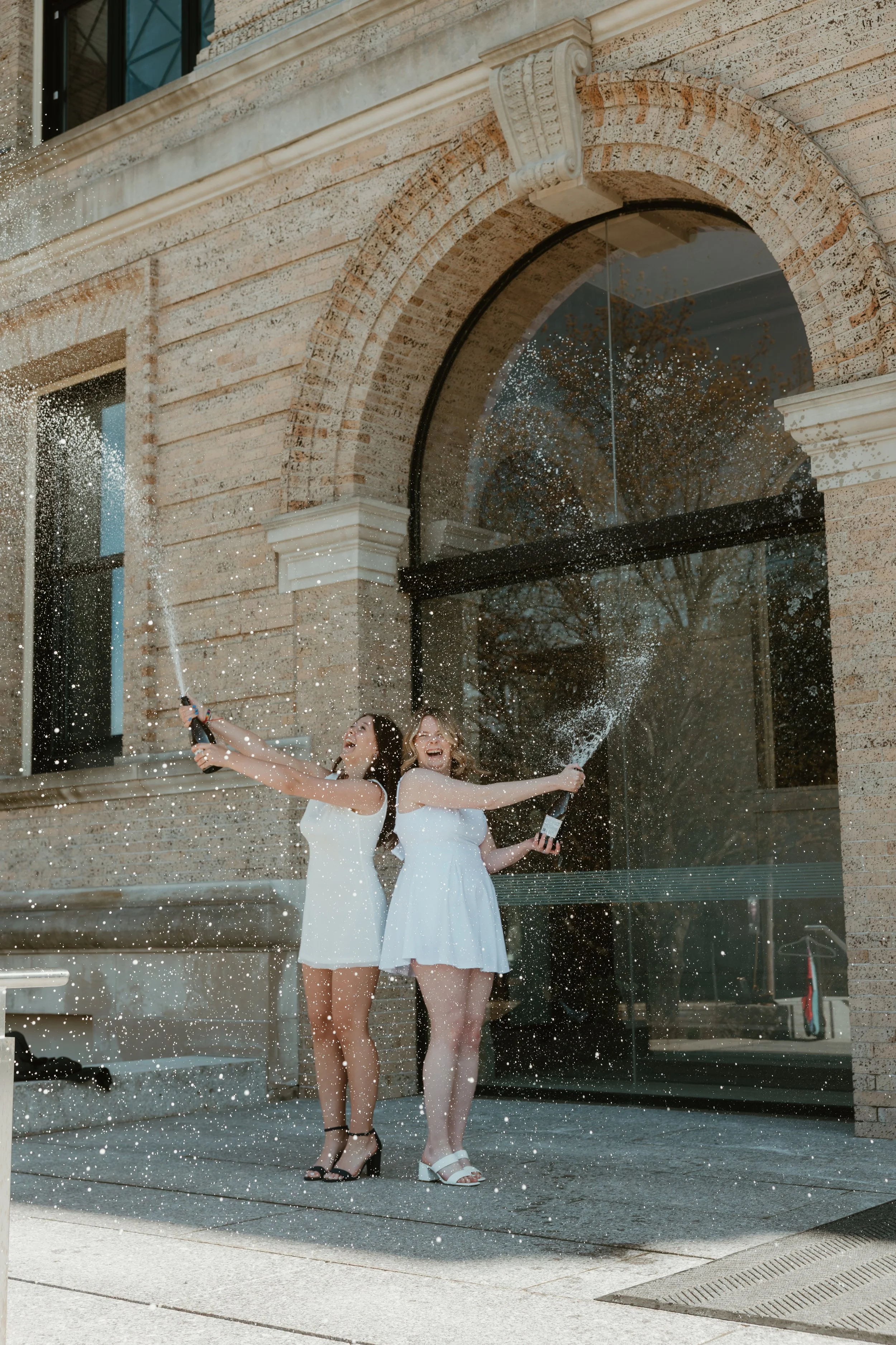 Two women in white dresses celebrating outside a brick building with large arched window, spraying champagne.