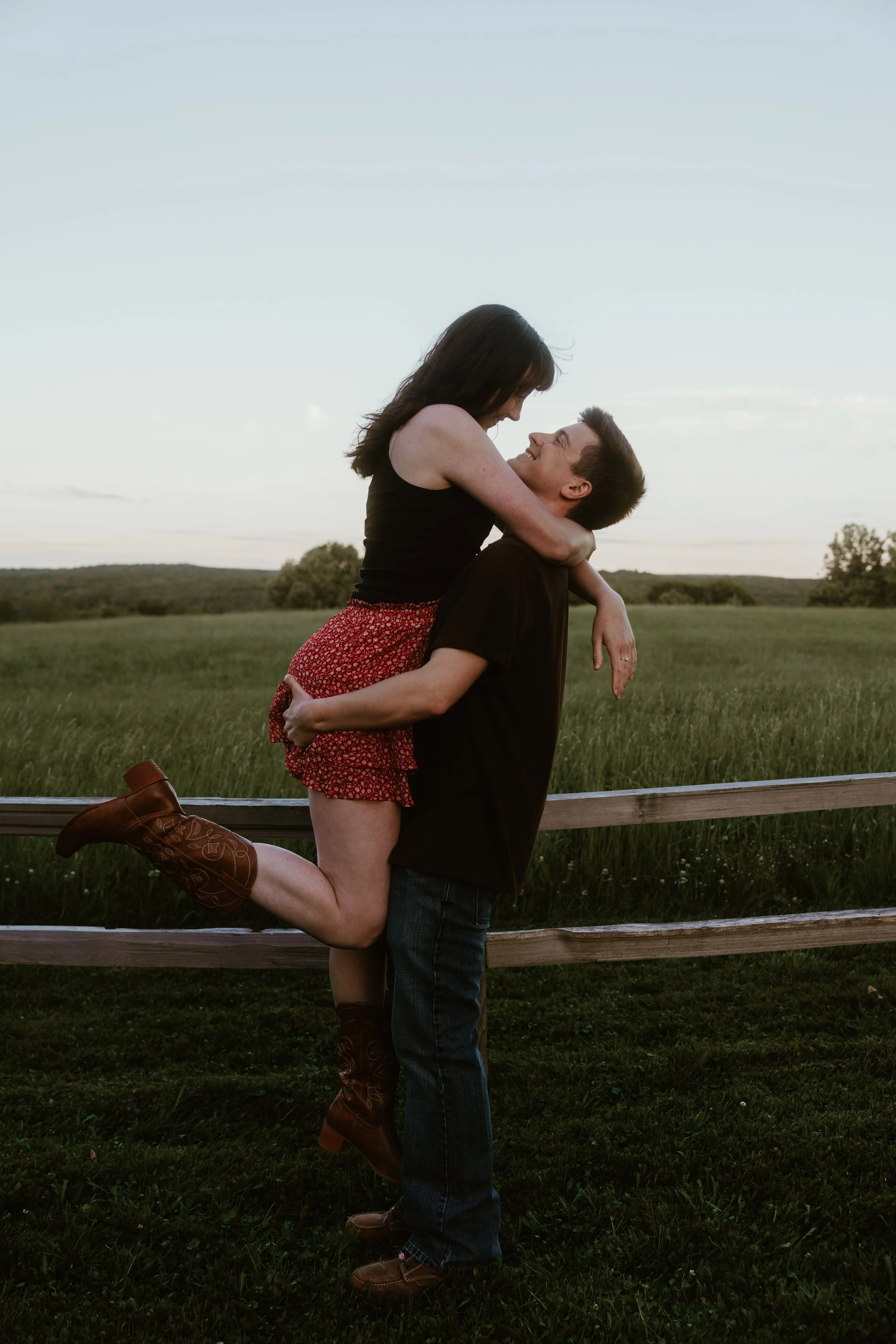 A young woman in brown cowboy boots, a black top, and a red skirt with white polka dots is being lifted by a young man in a black shirt and jeans outside. They are standing near a wooden fence with green fields and trees in the background, under a cl