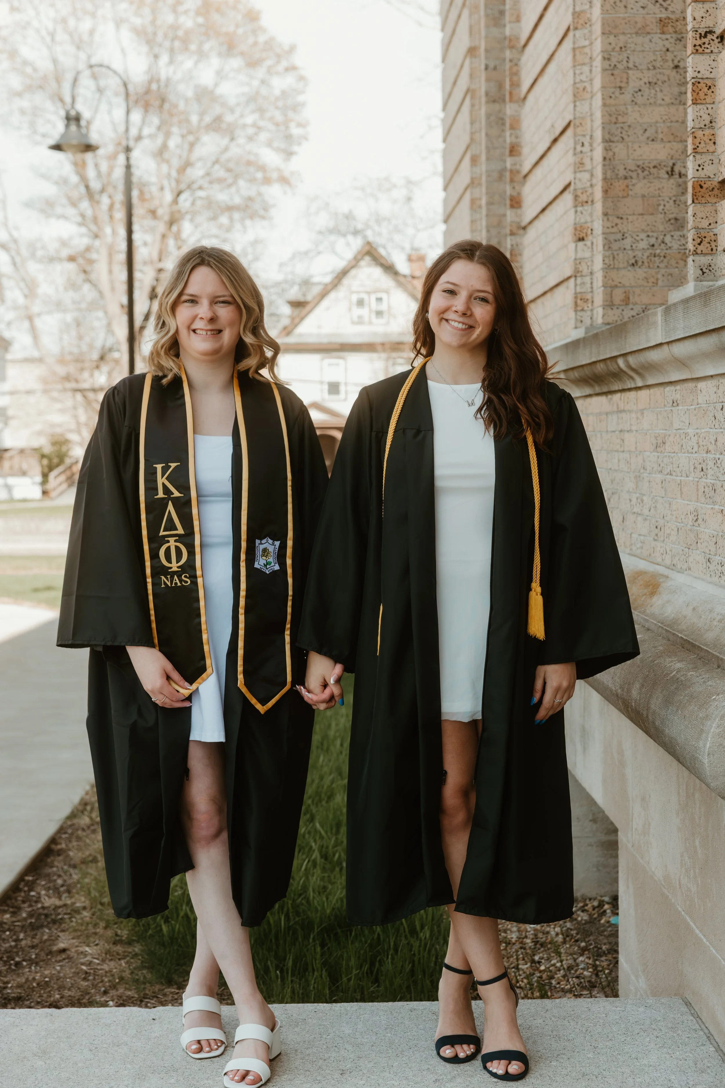 Two young women in graduation caps and gowns holding hands and smiling outdoors, one wearing white sandals and the other black heels, standing near a brick building with trees and a house in the background.