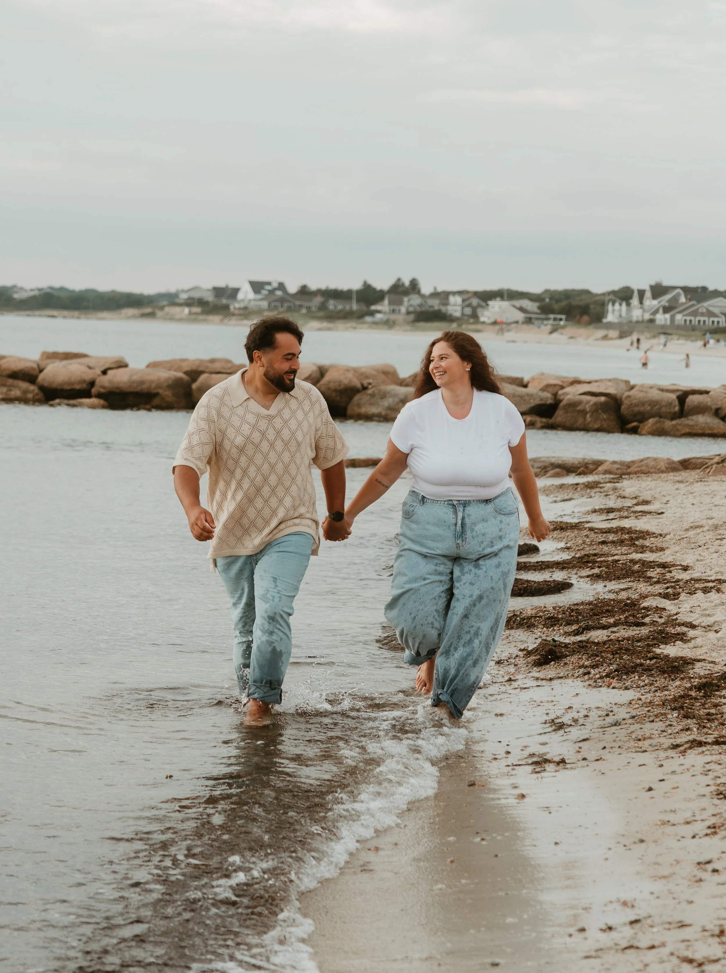 A couple holding hands and walking along the beach near the water, with houses in the background and rocks behind them.
