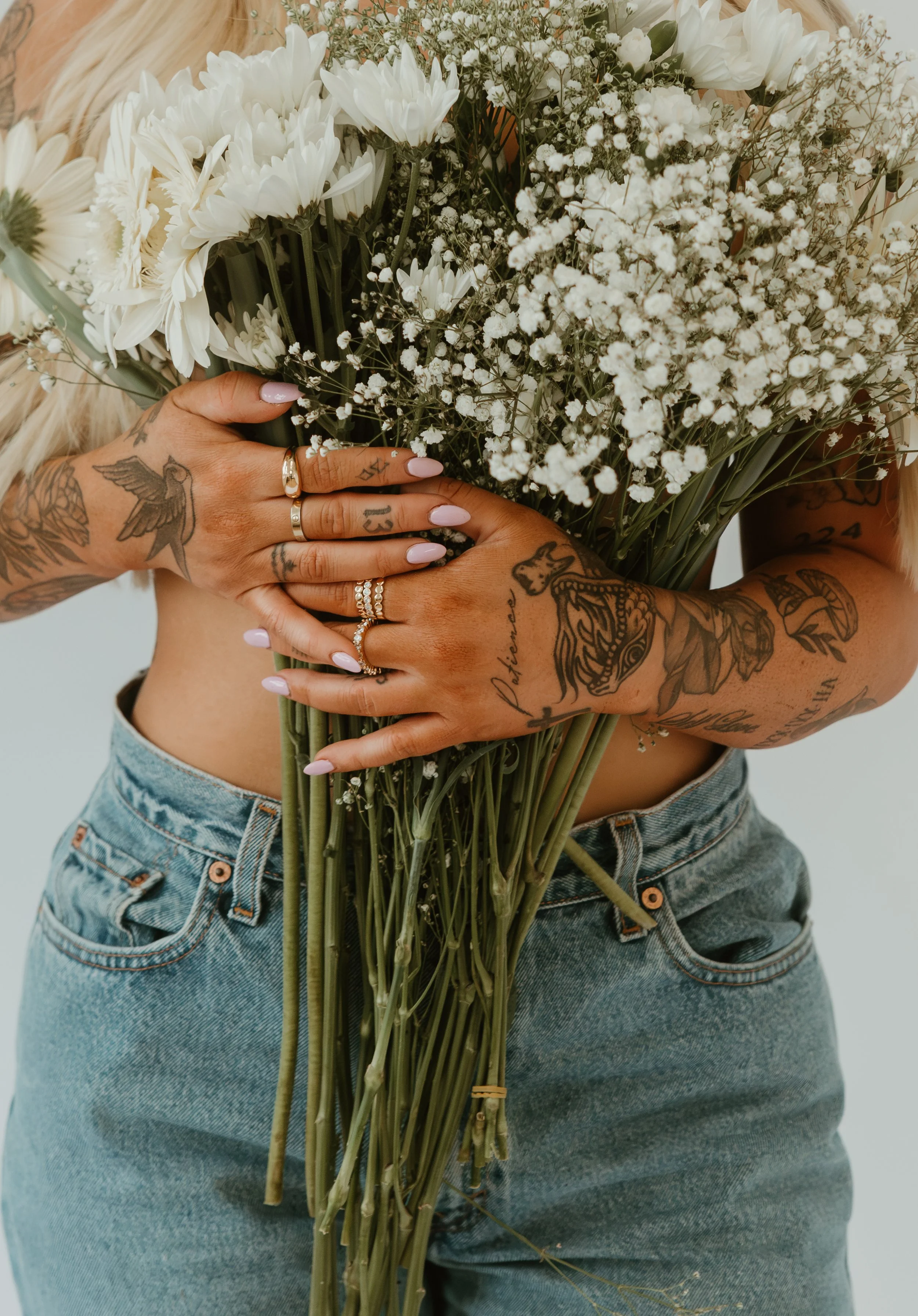 A woman with tattoos on her arms and hands holding a bunch of white flowers, including daisies and baby's breath, against a plain background. She is wearing rings and has painted nails, with her midriff and jeans visible.