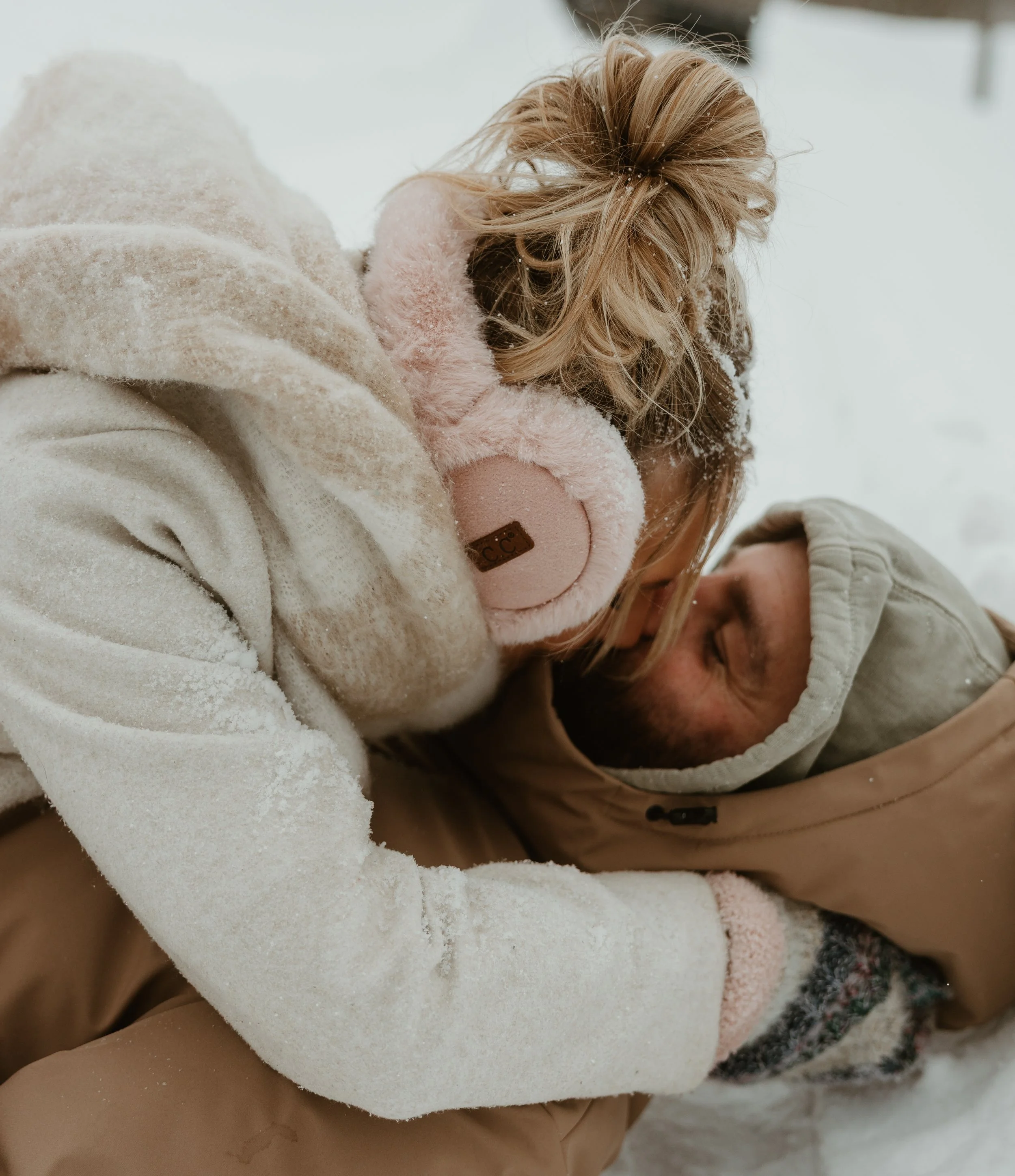 A person wearing beige clothing with a white fluffy sleeve, lying on their side in the snow during winter, with snowflakes on their hair and clothing, and another person's face partially visible in the foreground.
