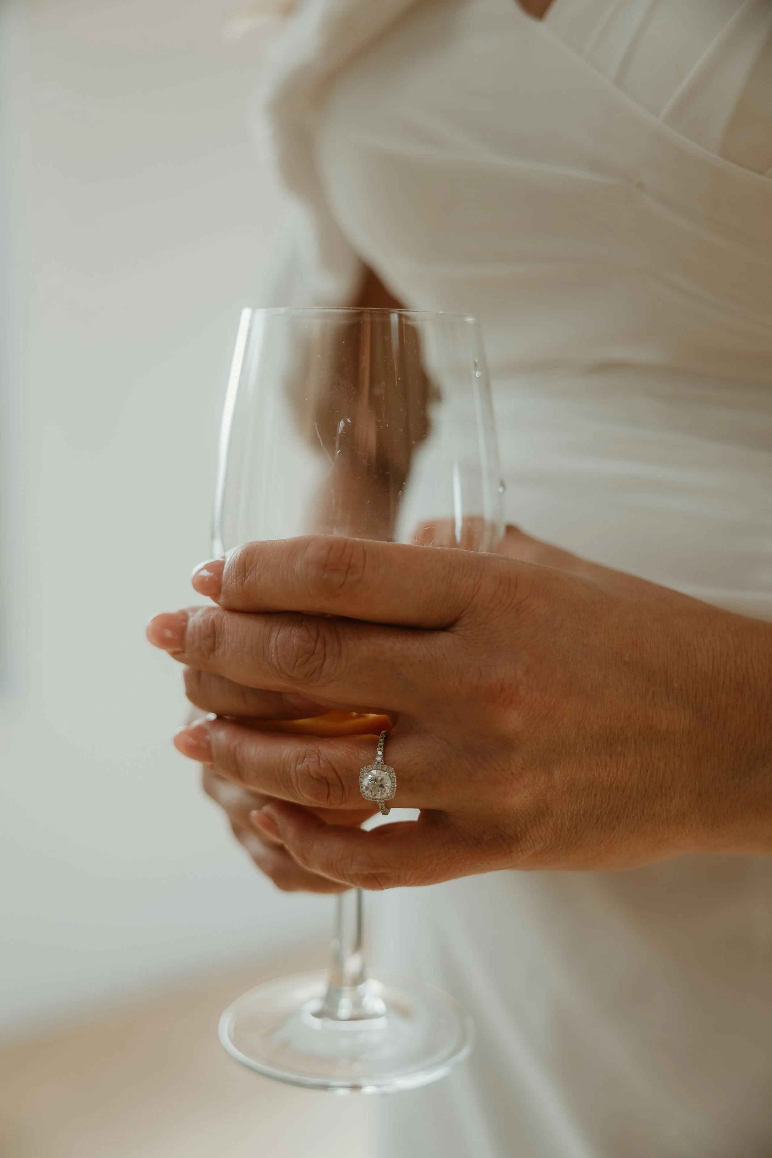 A person holding a wine glass in their hand. The person is wearing a white dress with a ring on their ring finger. The background is plain and light-colored.